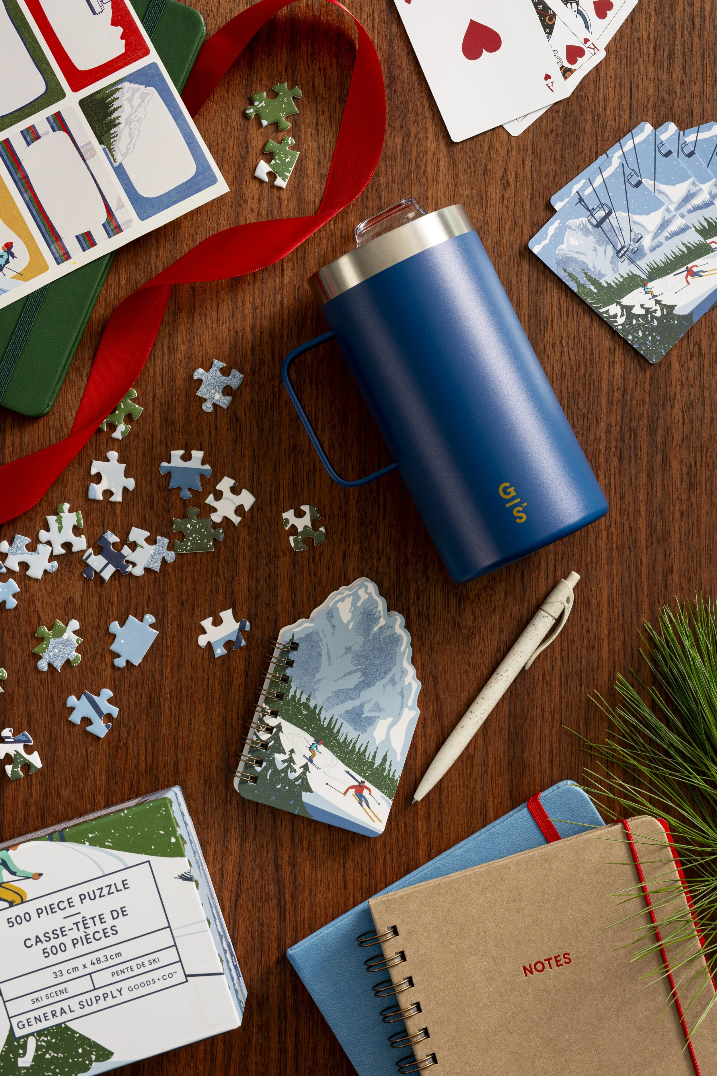 Various winter-themed items on a wooden table, including puzzle pieces, notebooks labeled 'Notes,' playing cards, a thermos, a pen, a small puzzle box, and decorative ribbons and greenery.