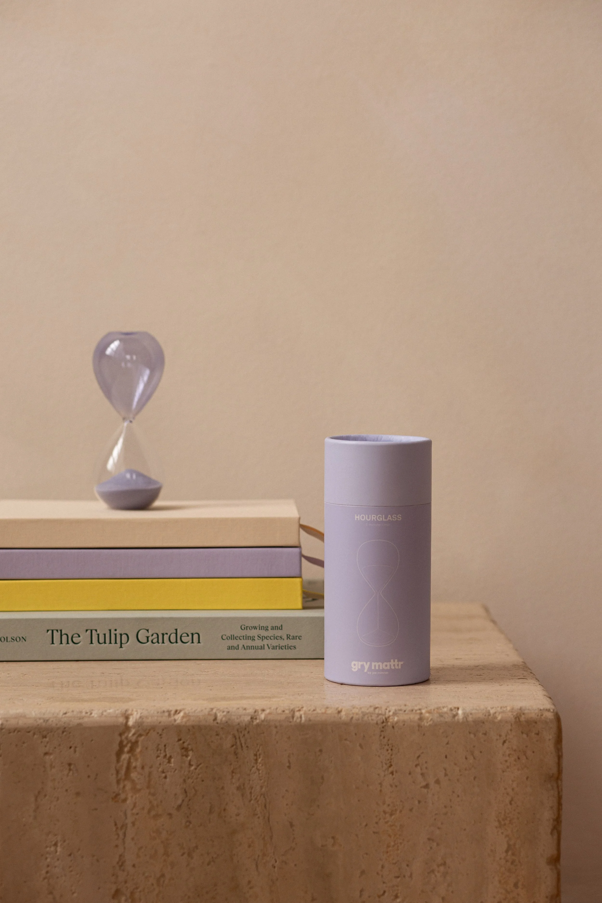 A white cylindrical object labeled 'gry matt' on a wooden surface, with a blue book titled 'The Tulip Garden' and a paperweight with a light-colored balloon design on top of the book, and a partially visible purple box underneath the book.