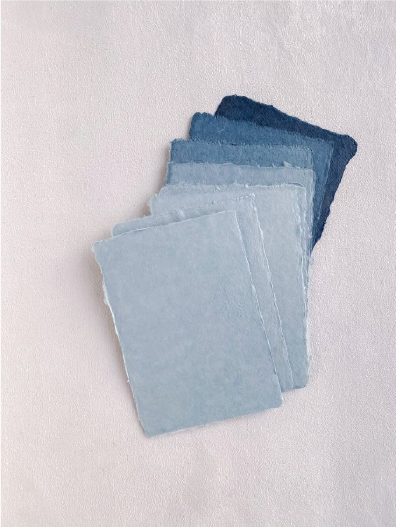 Stack of five watercolor paper sheets in shades of blue, arranged from dark to light, on a light-colored surface.