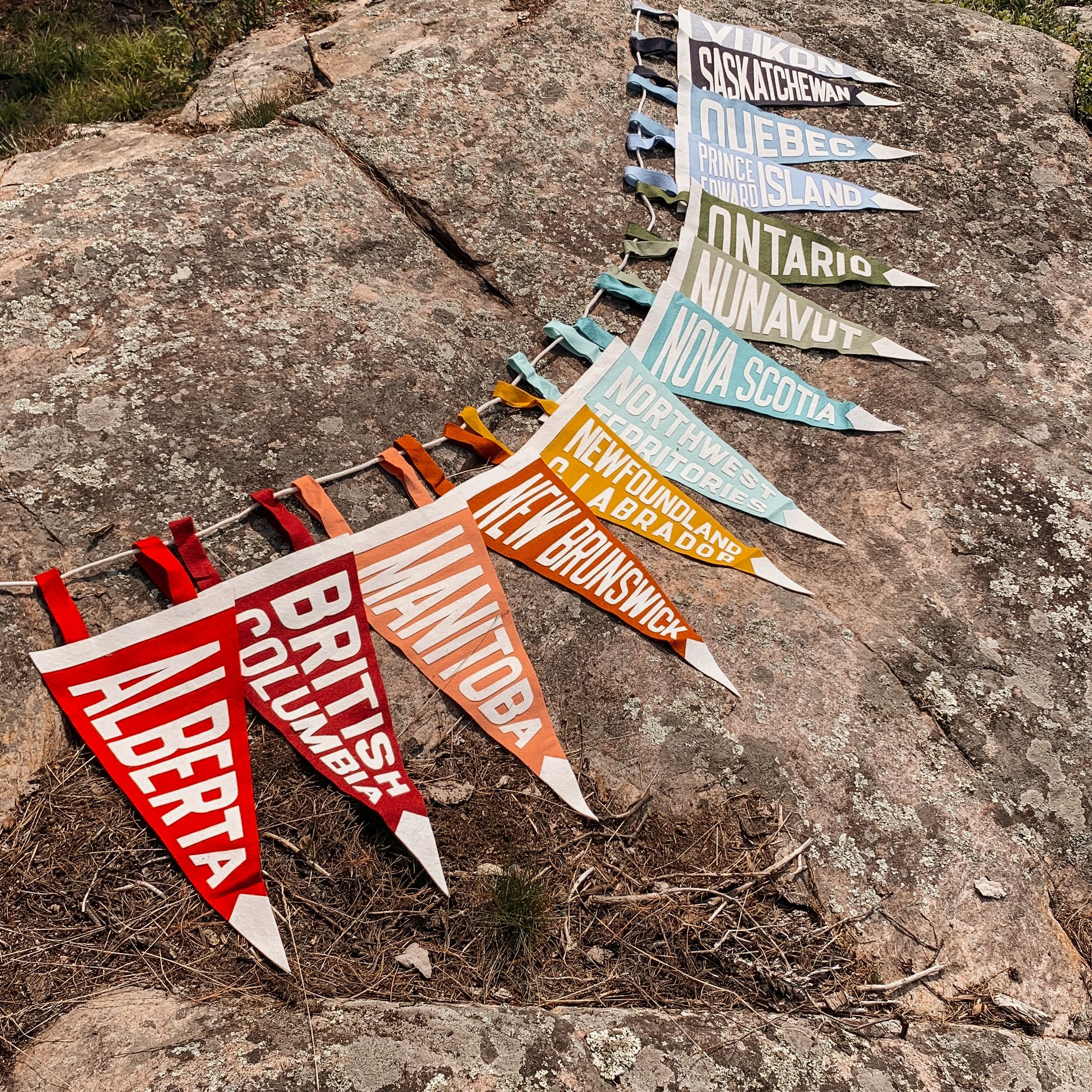 Colorful festival or parade flags with names of Canadian provinces, laid on a large stone surface outdoors.