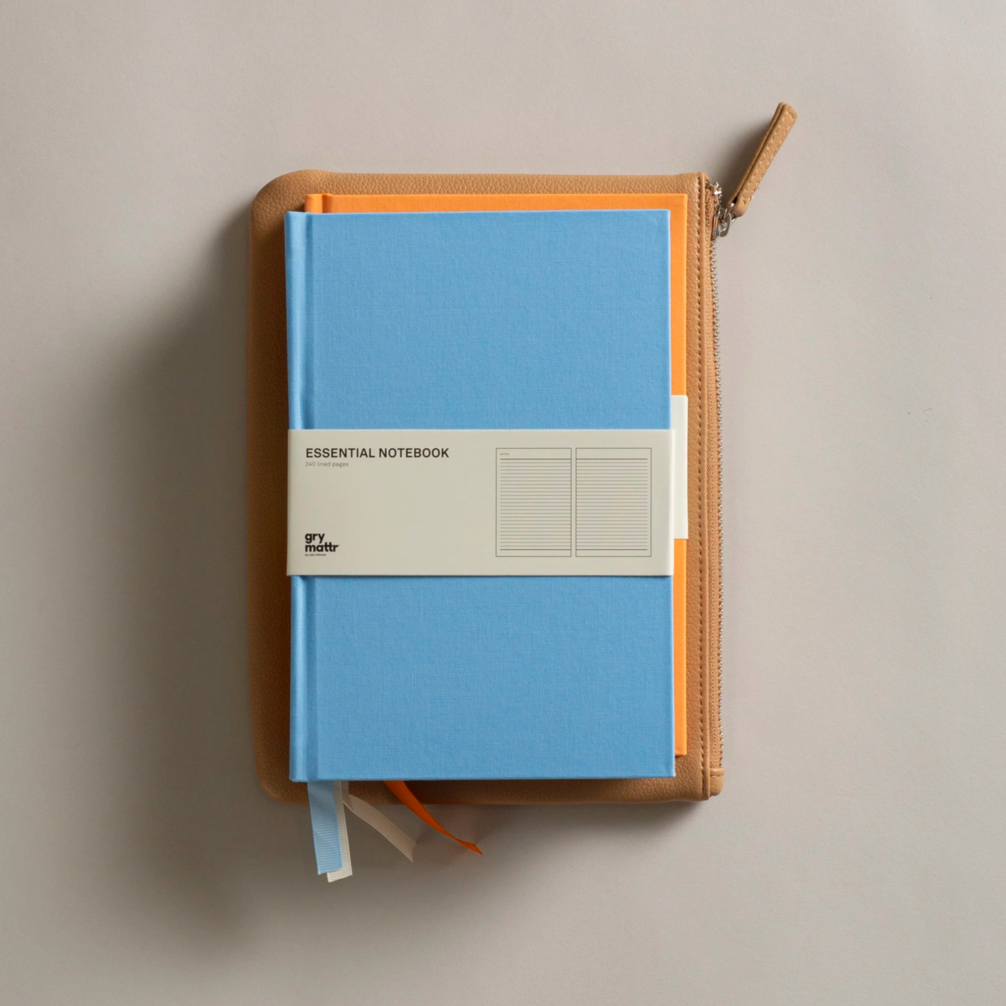 A tan textured zippered case with an orange zipper pull, partially unzipped, containing a light blue notebook labeled 'ESSENTIAL NOTEBOOK' in white, with a white band at the bottom labeled 'gry matrr' and a blue ribbon bookmark. The case is on a beige surface.