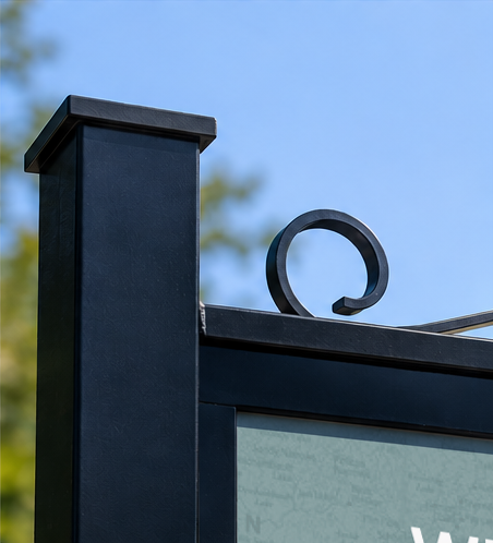 Close-up of a black metal sign or frame with a clear blue sky and blurred trees in the background.