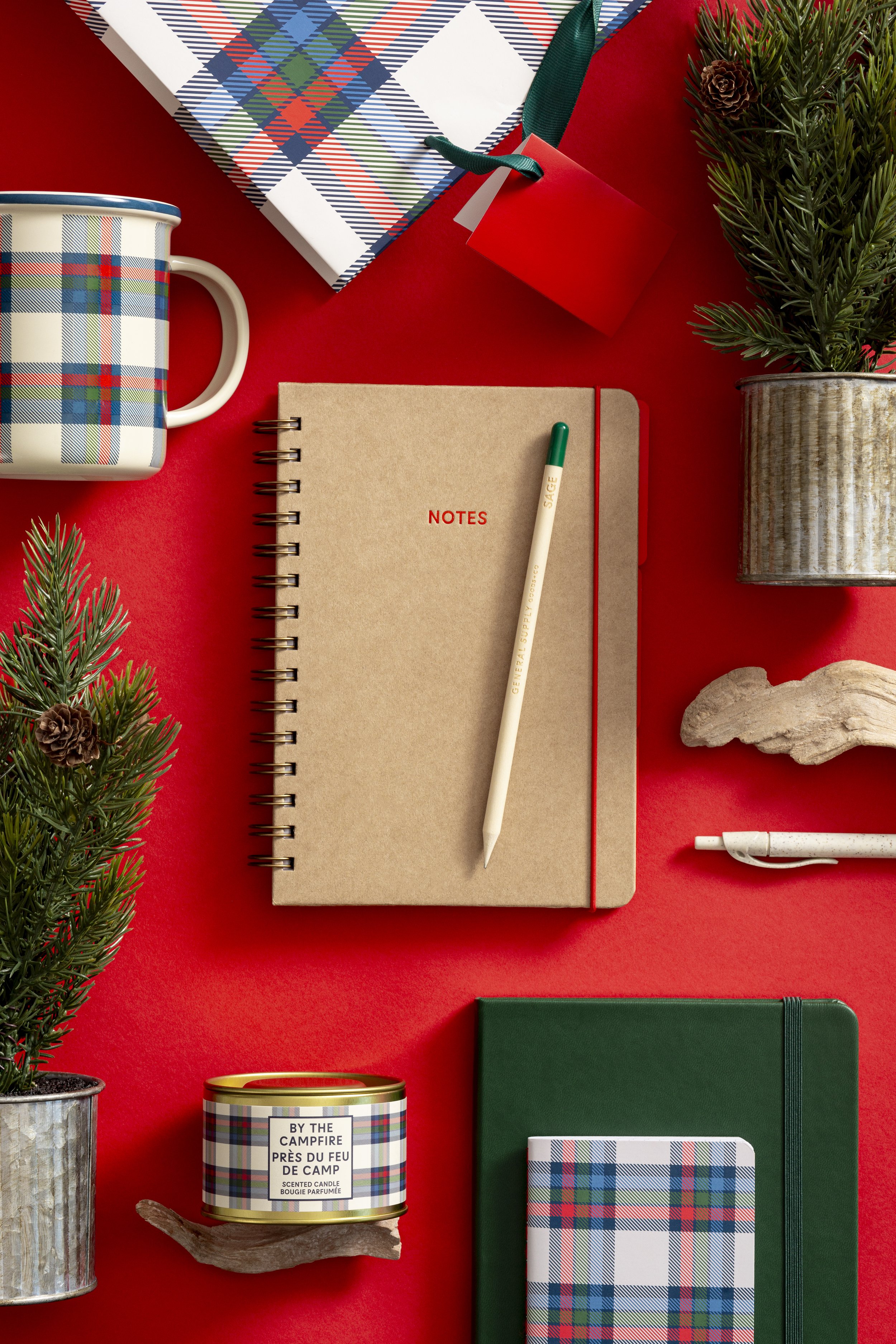 Flat lay of holiday-themed stationery and decorations on a red background, including a plaid gift bag, a notebook labeled 'NOTES', a white pen, a green notebook, a plaid patterned notebook, a plaid candle, a tin of candle labeled 'BY THE CAMPFIRE', a small potted pine branch with cones, and decorative rocks.