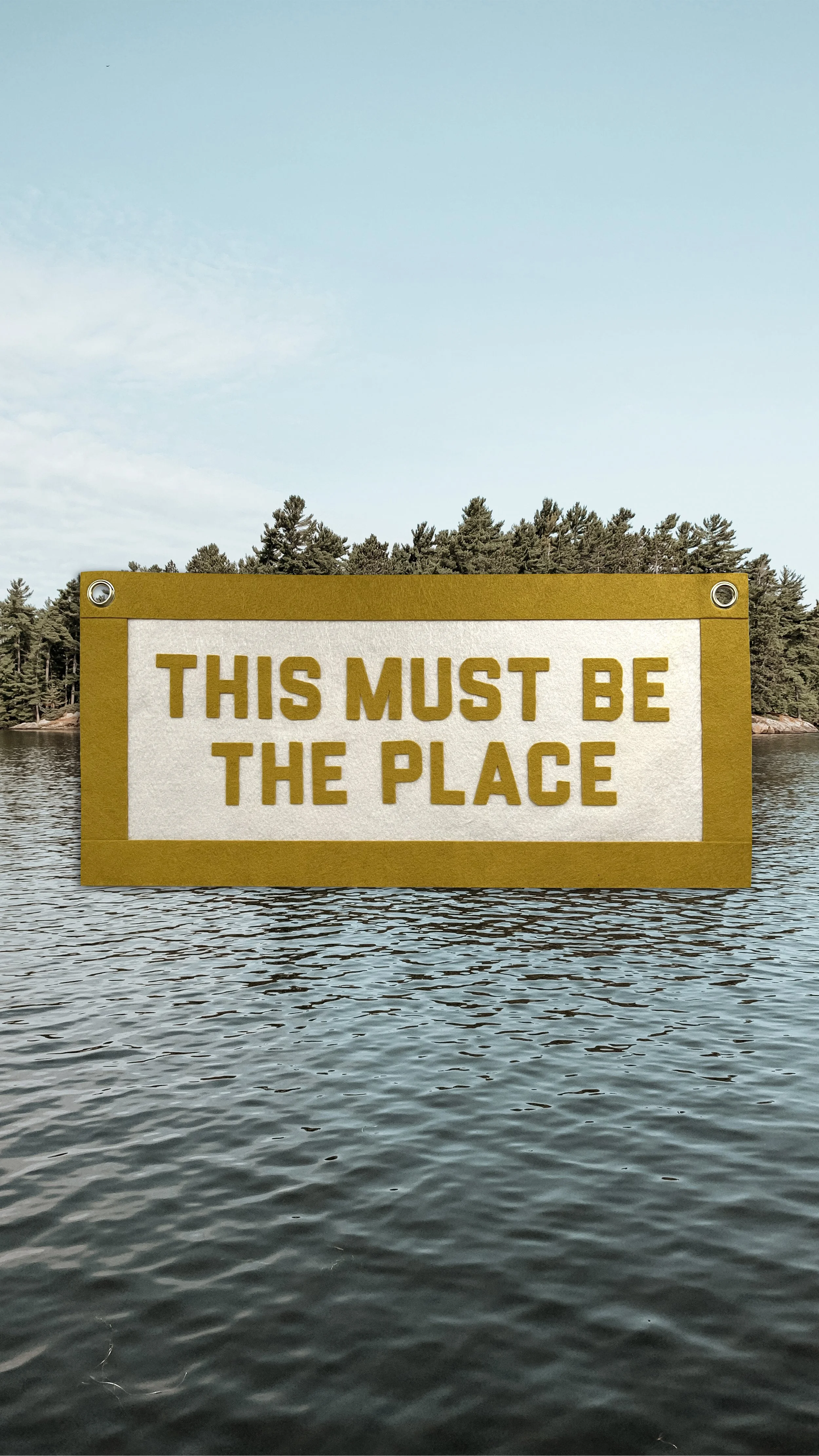 A floating sign that reads "This Must Be The Place" on water with trees in the background under a partly cloudy sky.