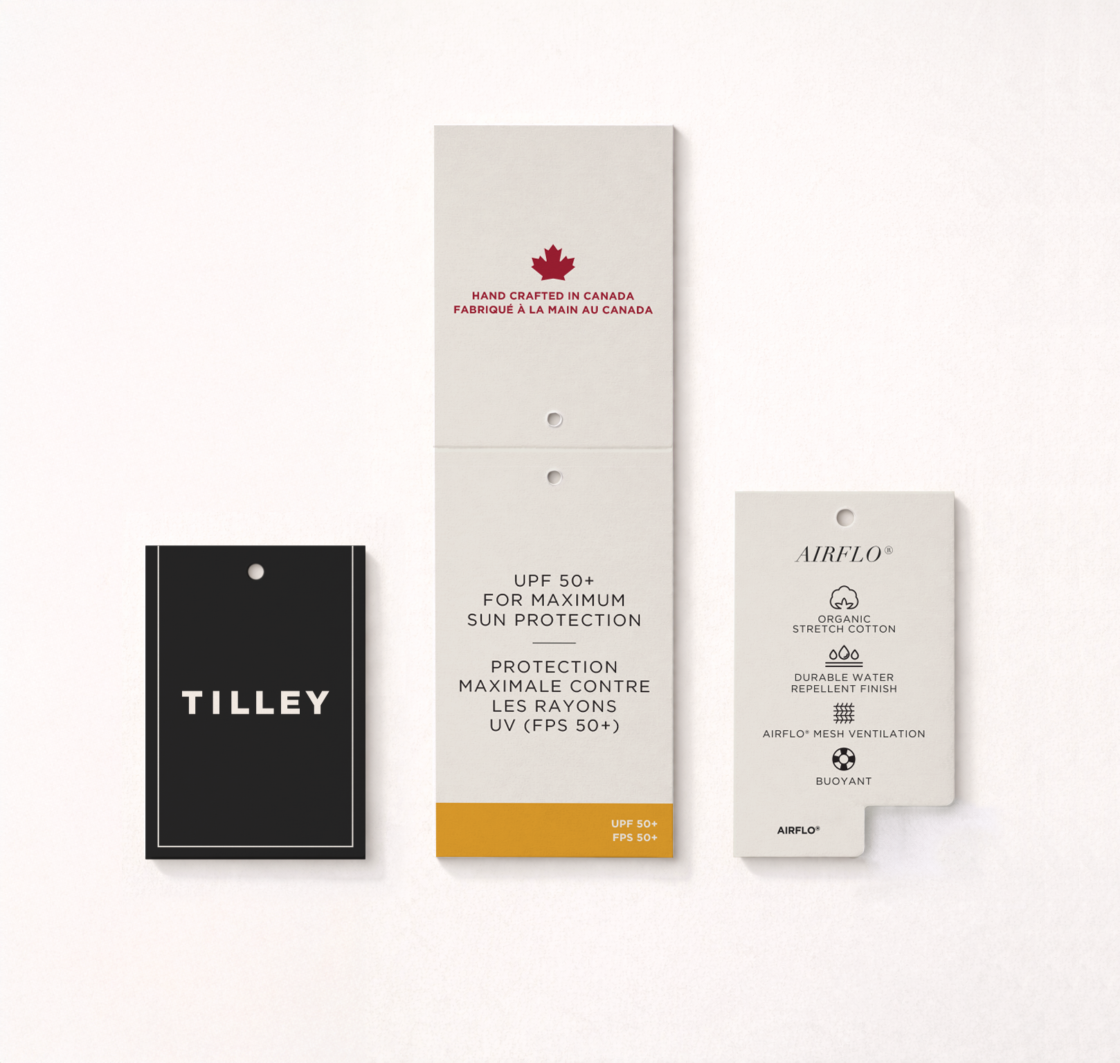 Three product tags for sunscreen and skincare products, including a black tag labeled "TILLEY," a white tag with red maple leaf symbol and text in English and French, and a white tag with black text and icons describing skincare features like organic stretch cotton, water repellency, mesh ventilation, and buoyancy.