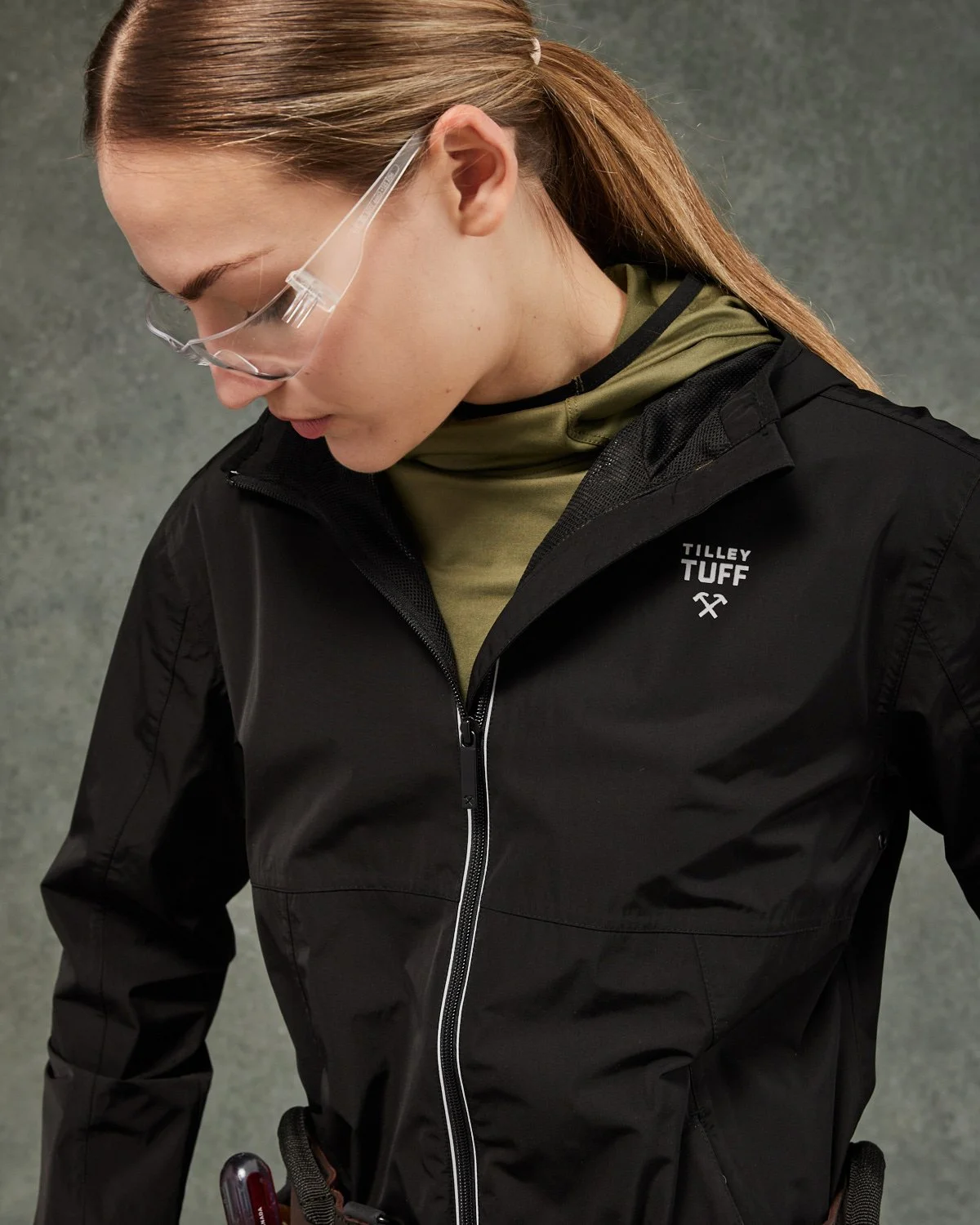 A woman wearing safety glasses, a black jacket with a zipper, and a khaki-colored top underneath. The jacket has a logo that says 'Tilley Tuff' with a small hammer and pickaxe symbol.