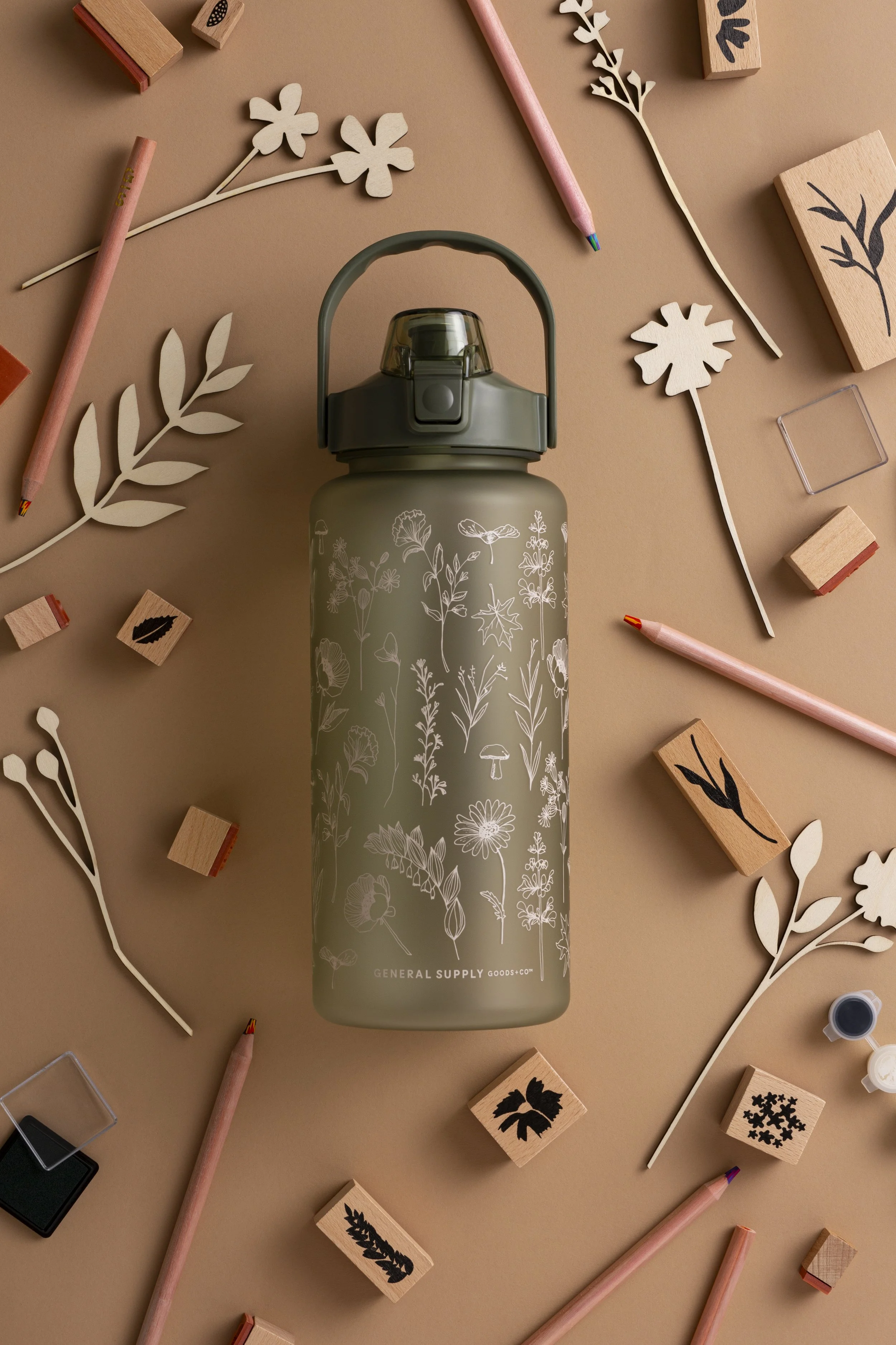 A beige water bottle with floral line art design and the text 'GENERAL SUPPLY GOODS + CO' surrounded by scattered wooden rubber stamps with botanical and nature-themed designs, and pink pencils on a beige surface.