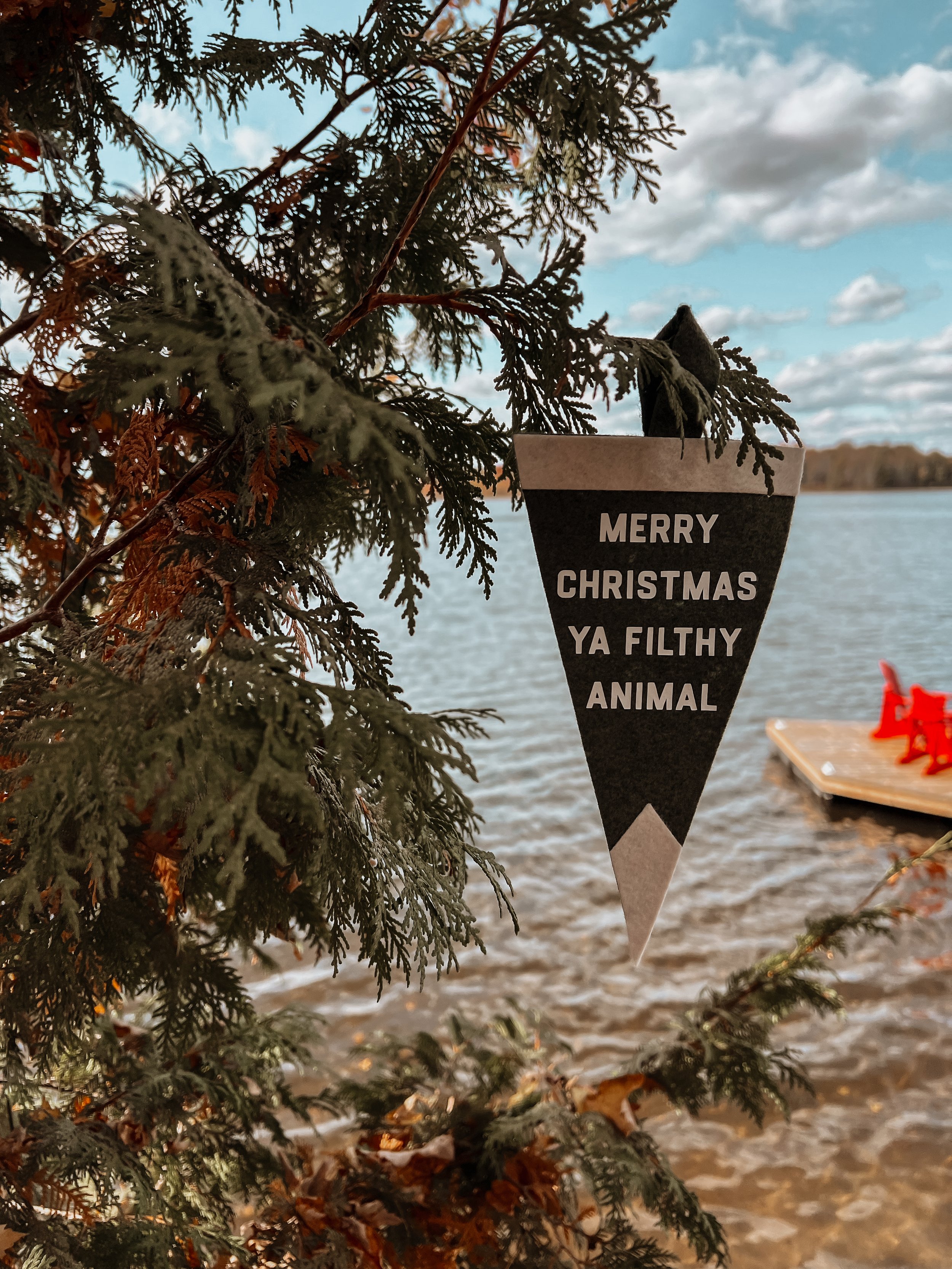 A Christmas tree branch with a black and white ornament that reads "Merry Christmas Ya Filthy Animal" hanging from it. In the background, a lake under a partly cloudy sky is visible.