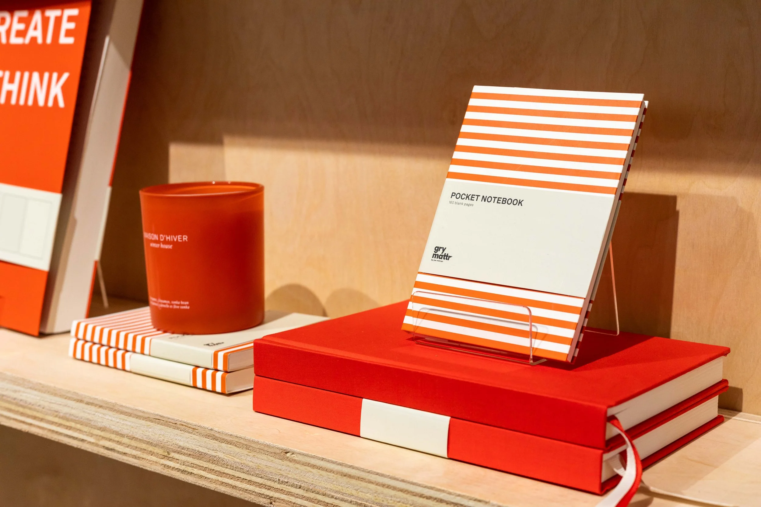 A display of various notebooks with red, white, and orange striped covers, arranged on a wooden shelf, including a pocket notebook titled 'groymattr' and a red cylindrical container.