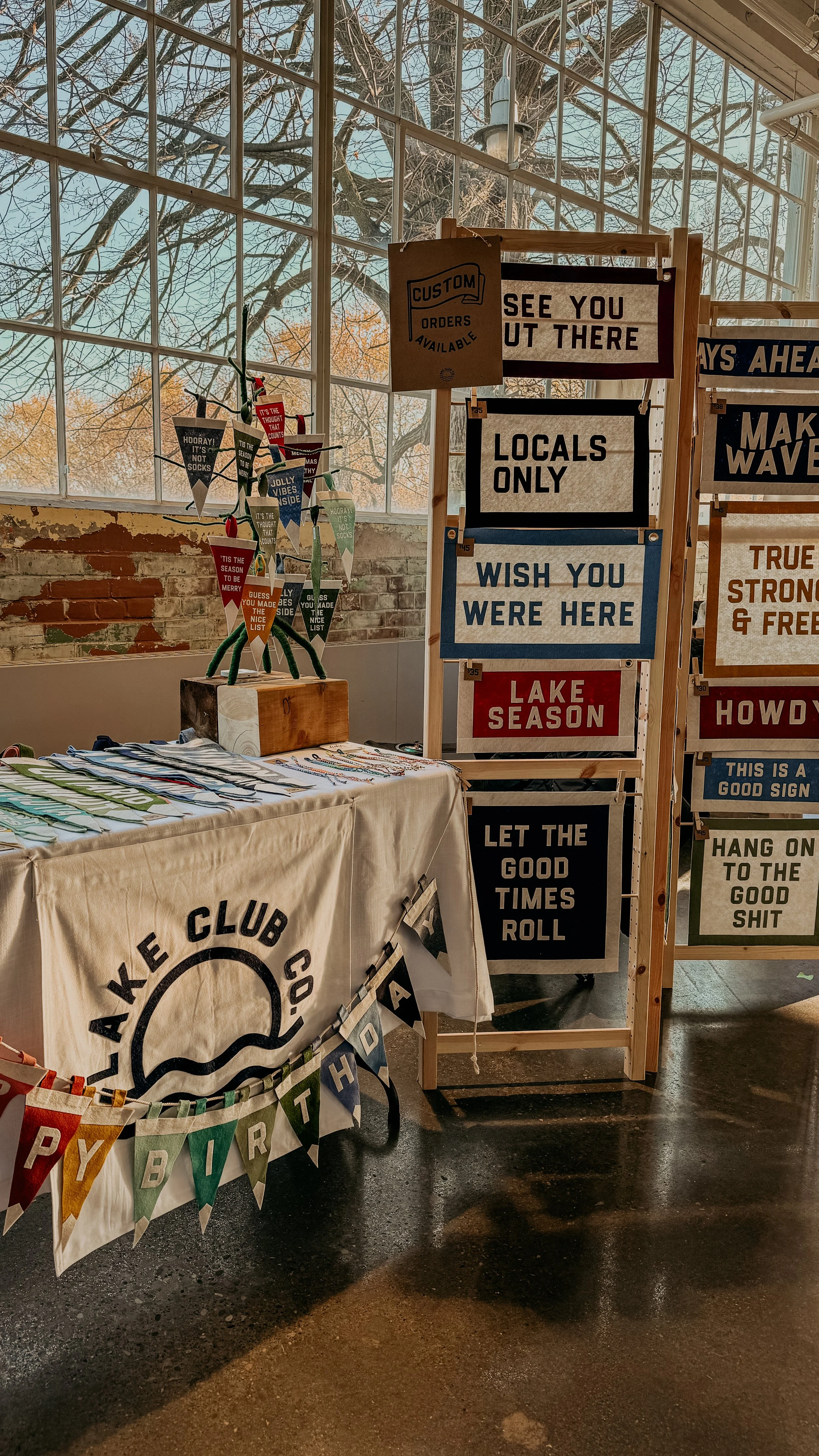 A booth with a beige tablecloth with black text and a logo, and a display board with various signs with slogans about local events, good times, and season's greetings. There are also small flags or banners hanging on the table and a small decorative 