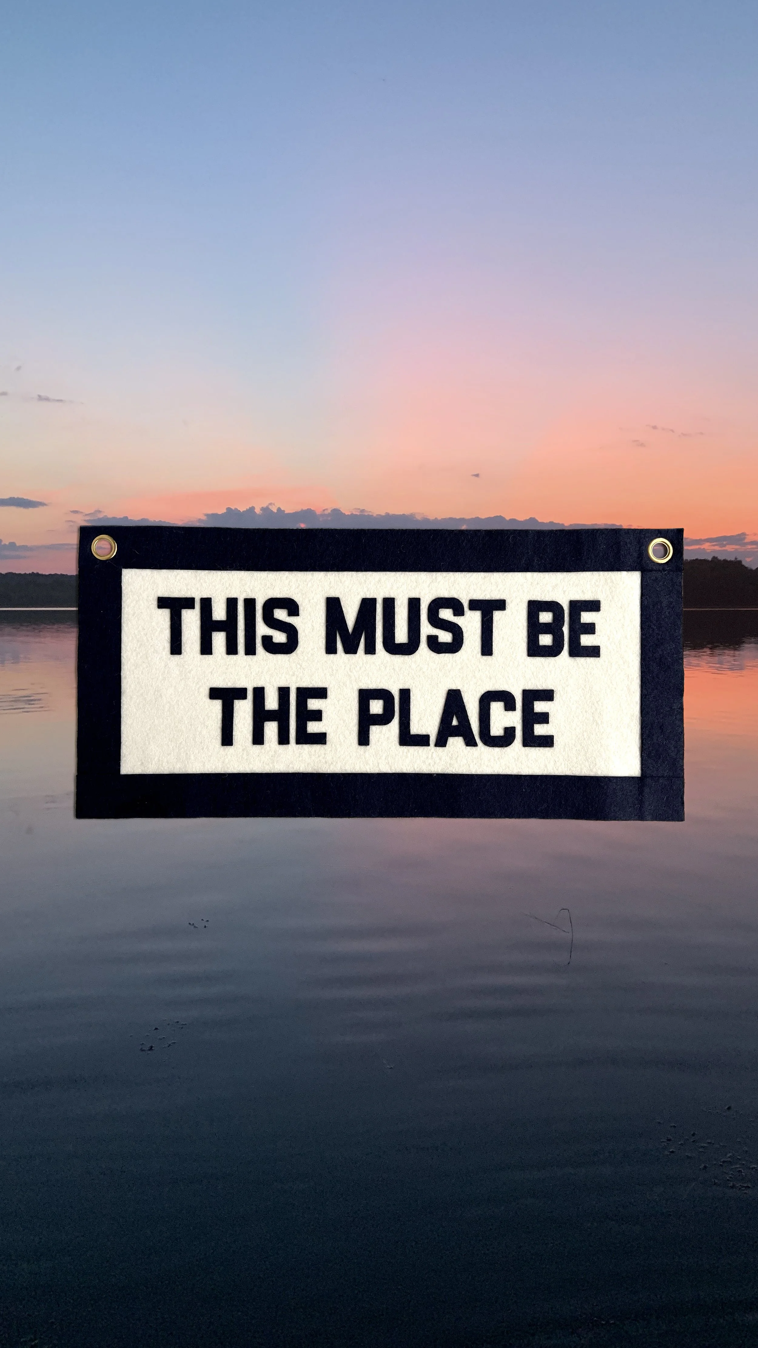 Sign with the text "This Must Be The Place" against a sunset over a calm lake.