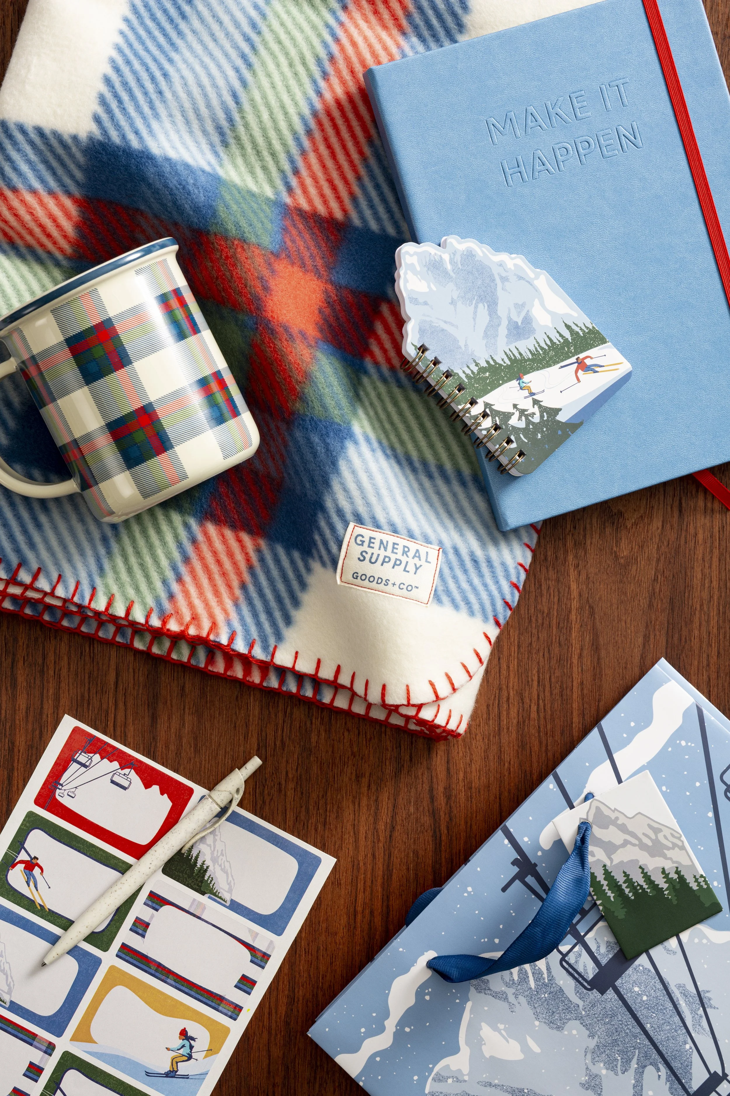 A holiday scene with colorful items including a plaid mug, a blue notebook with the phrase 'MAKE IT HAPPEN,' a small spiral notebook with a snow mountain graphic, and illustrated postcards featuring skiing scenes, snow mountains, and winter sports, all placed on a plaid blanket with red, blue, green, and white colors.