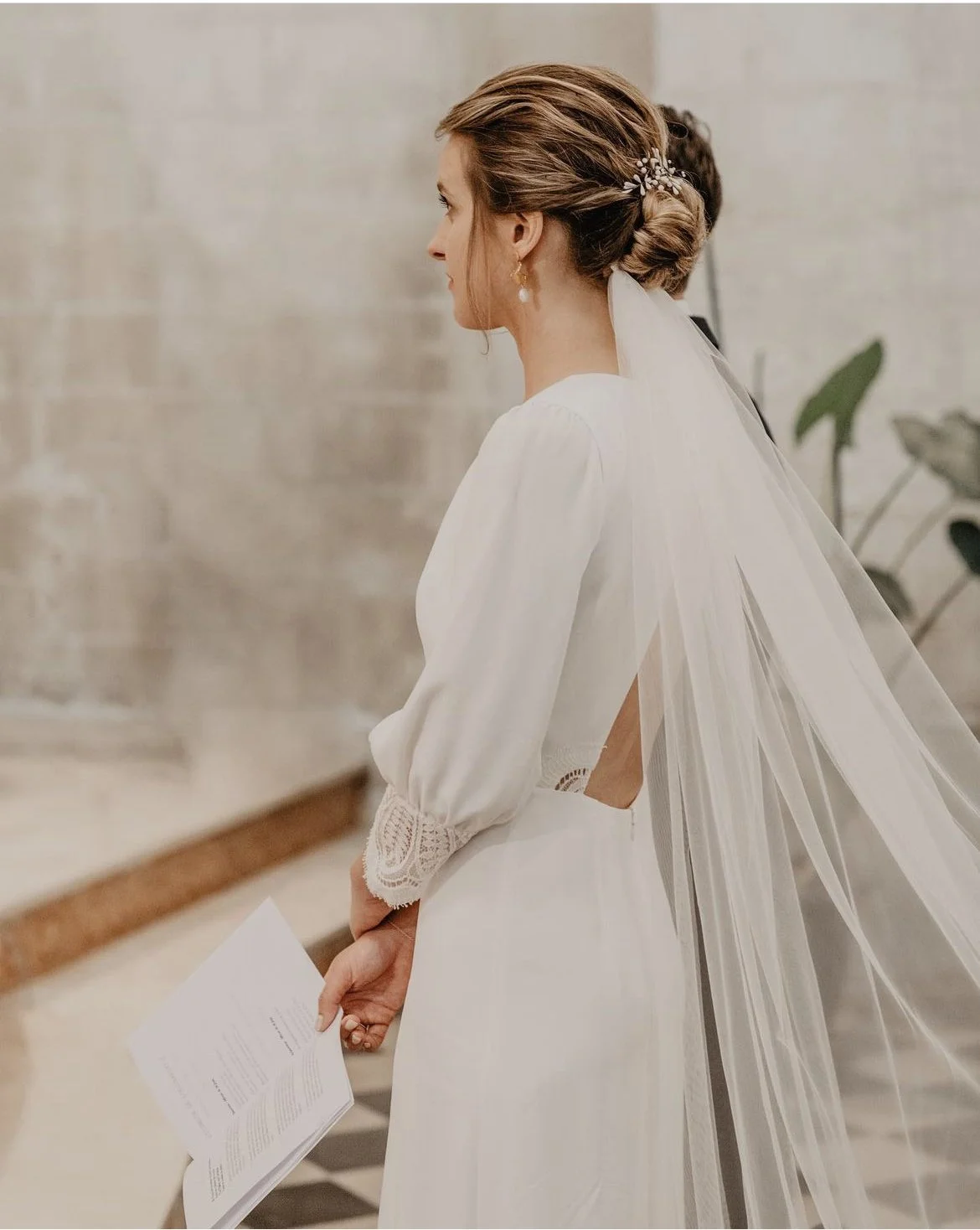 Une femme en robe de mariée blanche avec un voile, vue de profil, tenant un document dans la main.