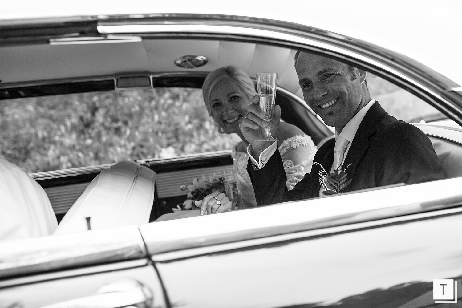 A black and white photo of a smiling couple in a car, the man holding a glass and the woman wearing a wedding dress.