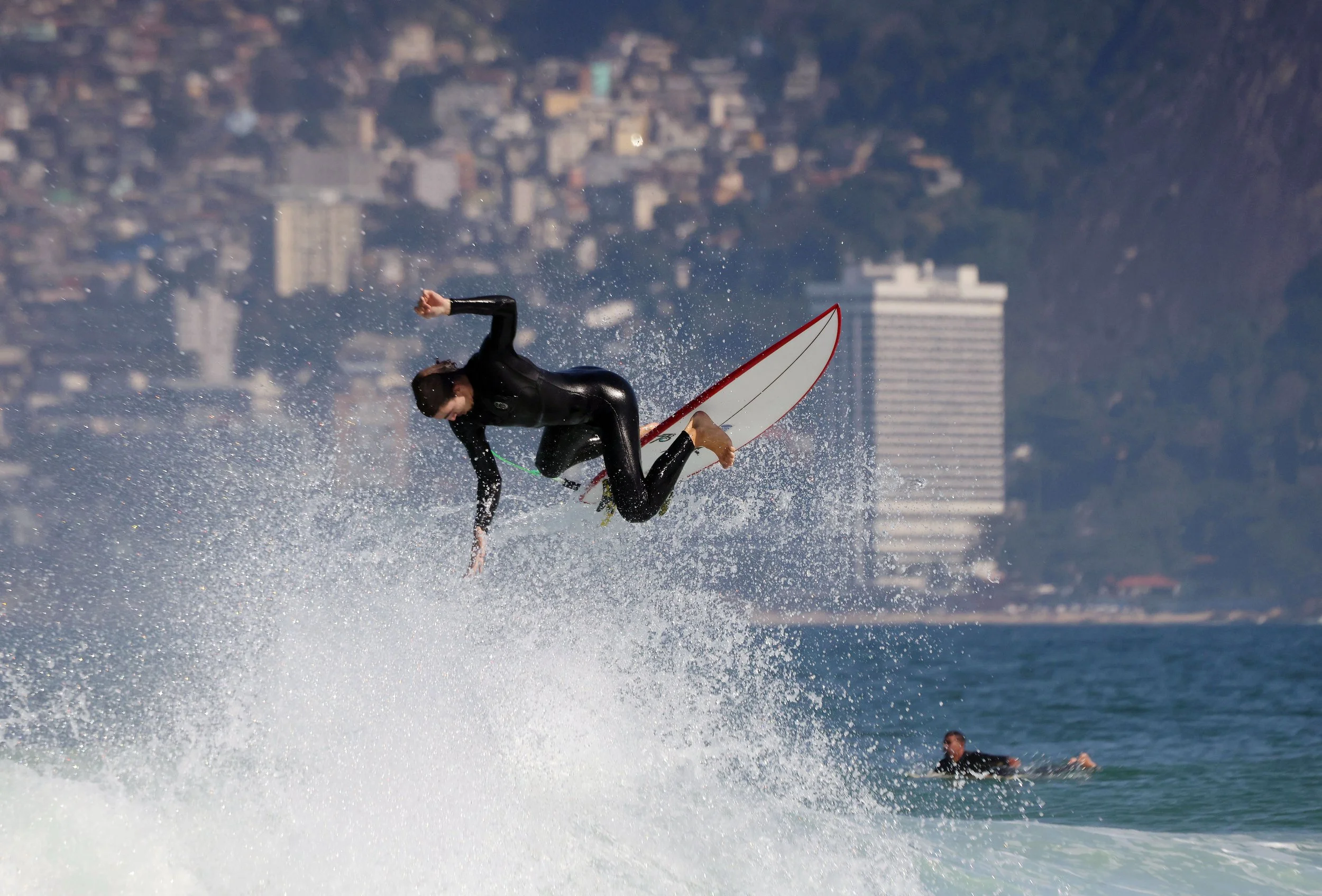 Surfer BENJAMIN D’OREY launches into the air at Arpoador, trying to hold onto his board during a dynamic maneuver, with Rio’s Vidigal neighborhood rising in the background.