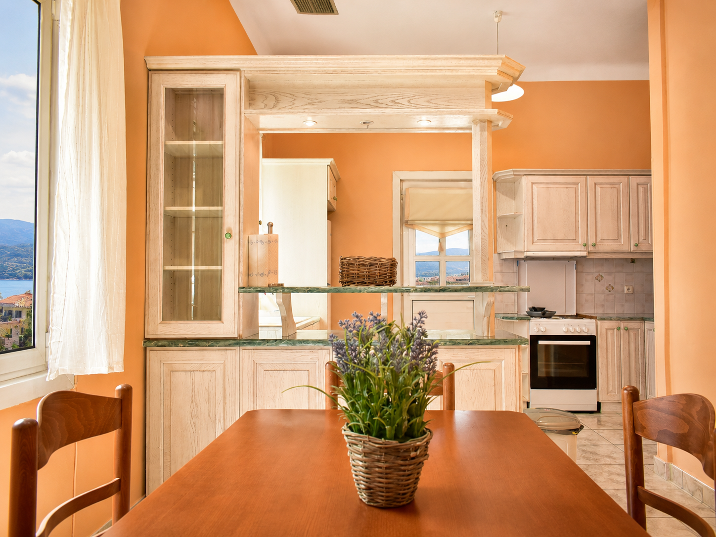 Dining area with a wooden table and chairs, a potted plant centerpiece, and a view into a kitchen with cream cabinets, a stove, and windows overlooking a landscape.