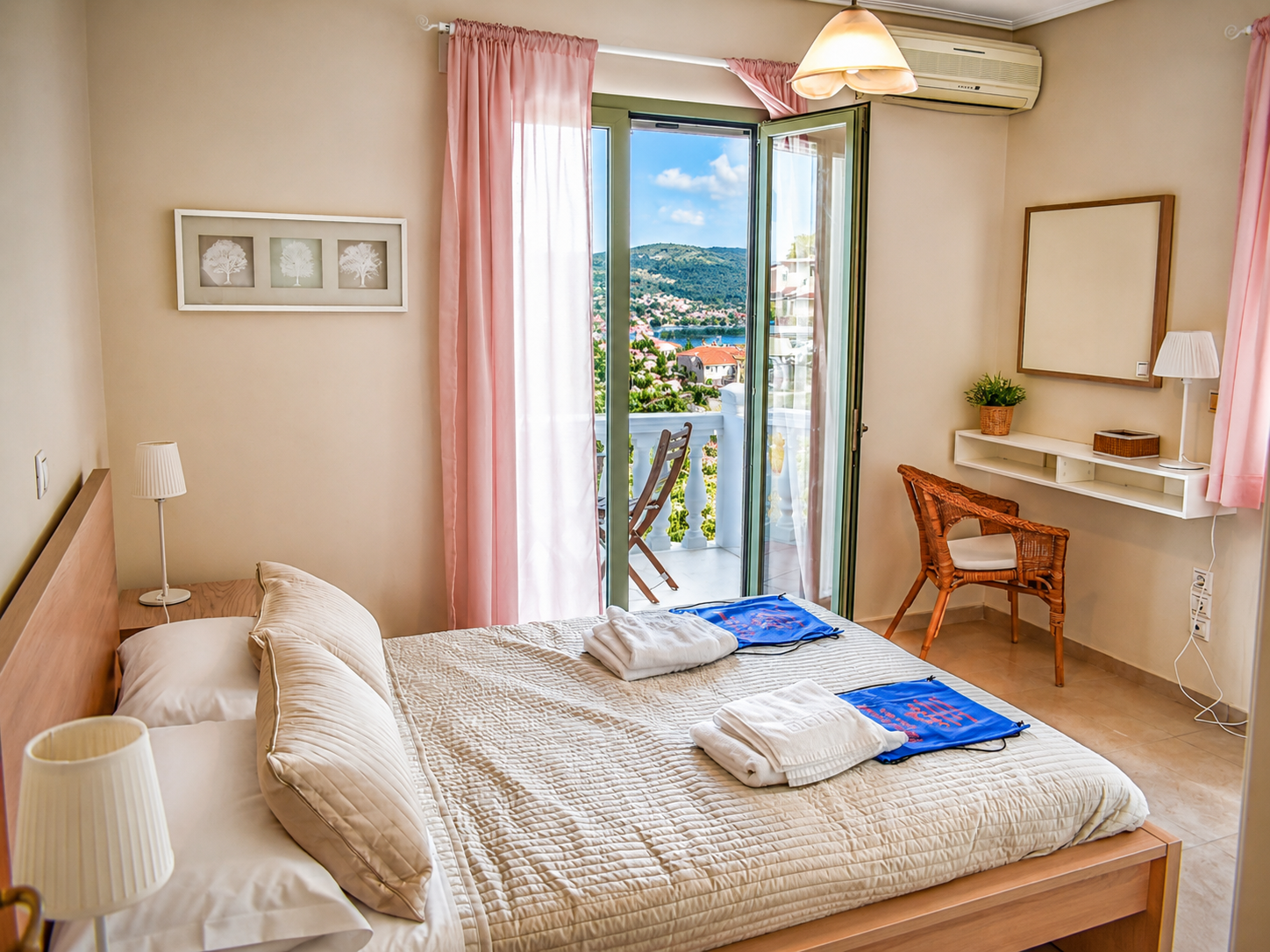 A cozy hotel bedroom with a double bed, pink curtains, a small balcony with a chair, and a view of mountains and houses outside.