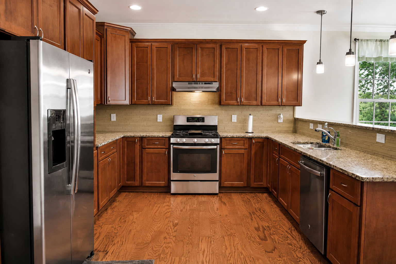 Kitchen with wooden cabinets, stainless steel refrigerator, oven, dishwasher, granite countertops, hardwood floor, window with green trees outside, and pendant lights.