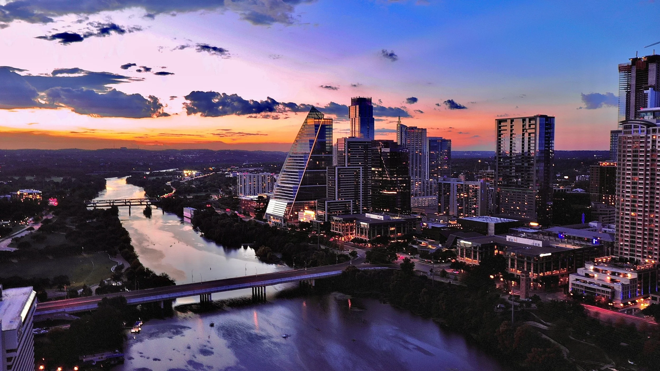 A city skyline at sunset with tall skyscrapers, a river running through the city, and a partly cloudy sky with purple and orange hues.