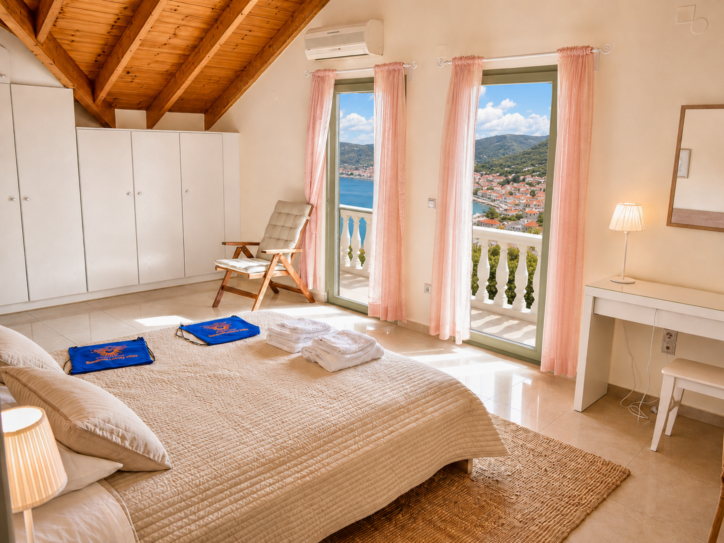 A bright bedroom with a large bed, pink curtains, and a view of a coastal town from a balcony. The room has a wooden ceiling, white cabinets, a beige chair, a white desk with a lamp, and folded towels on the bed.