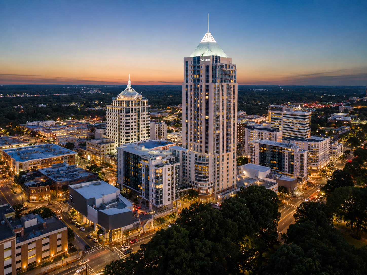 City skyline at dusk featuring tall modern high-rise buildings, illuminated with lights, with trees and streets in the foreground and a sunset sky in the background.