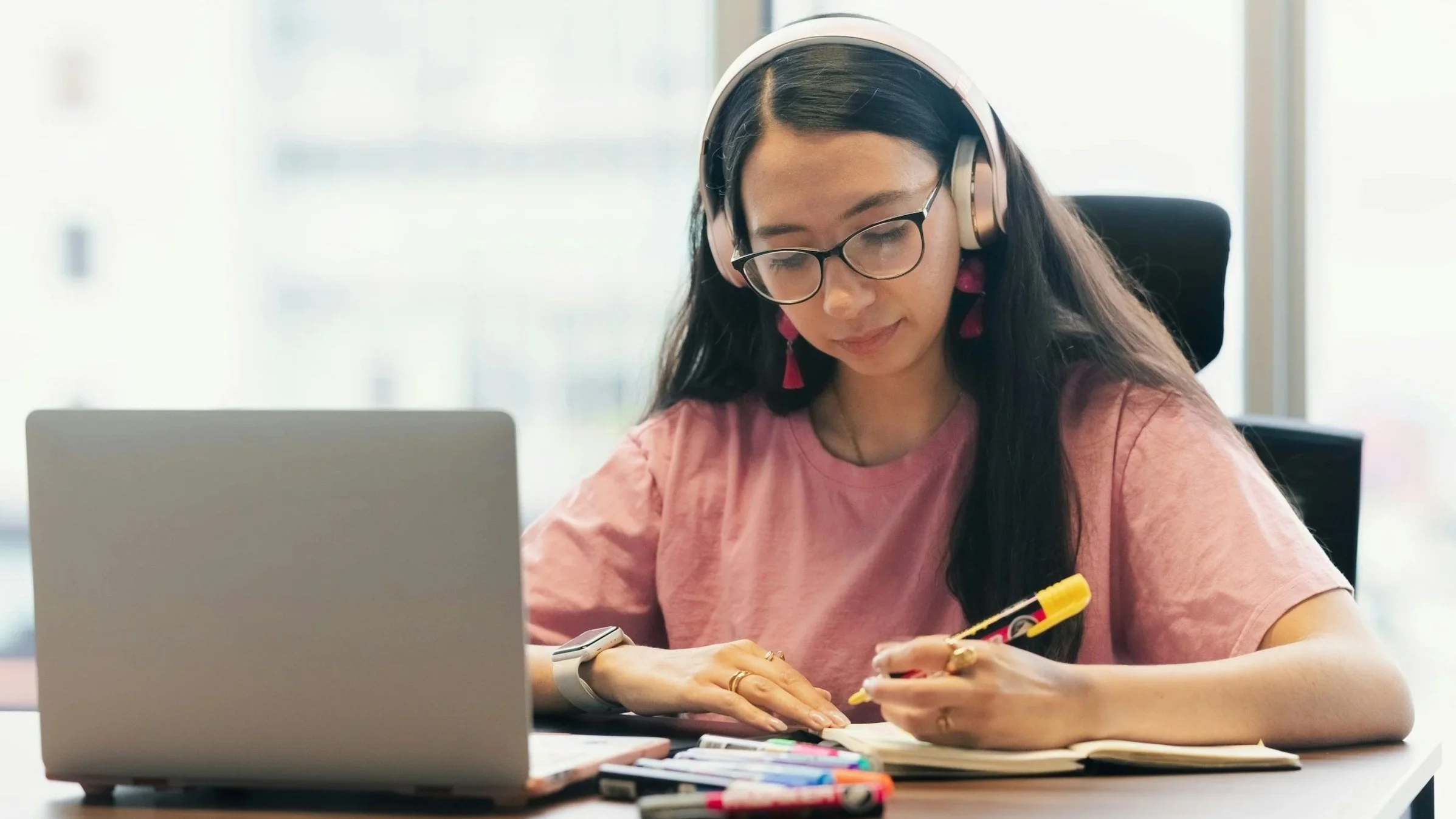 Young adult writing in a notebook while working on a laptop with headphones.