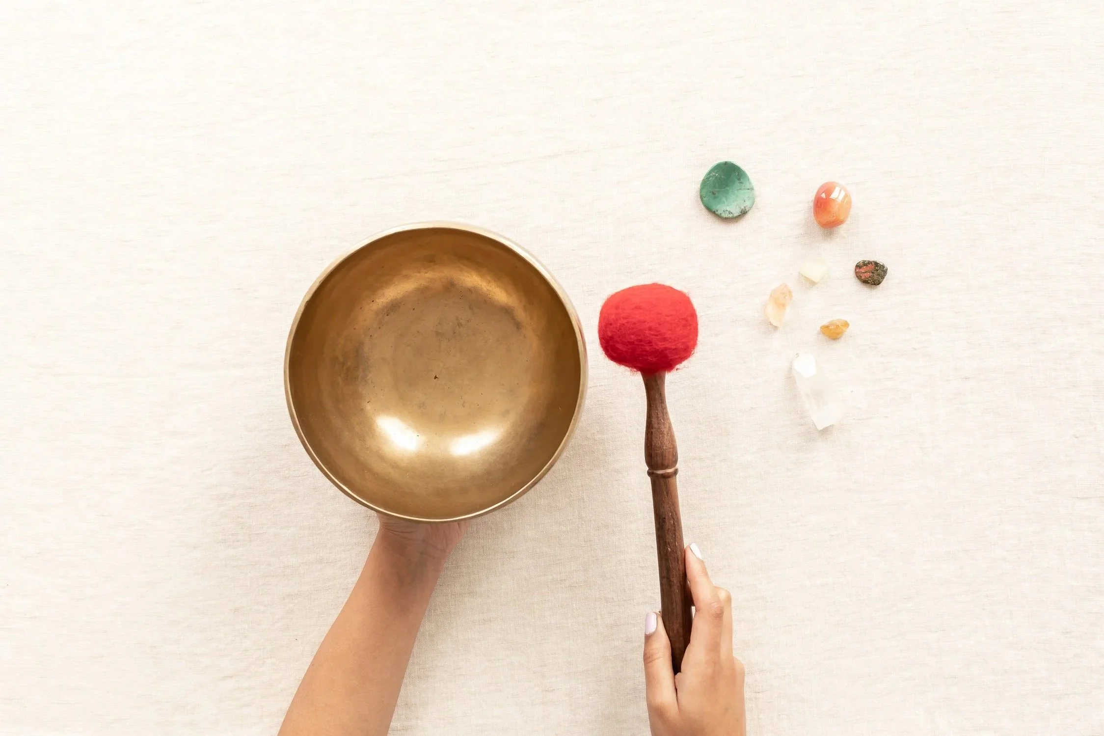 A hand holding a small, rounded brass singing bowl and another hand holding a red felt mallet. Several small colorful stones and crystals are arranged on a light-colored surface.