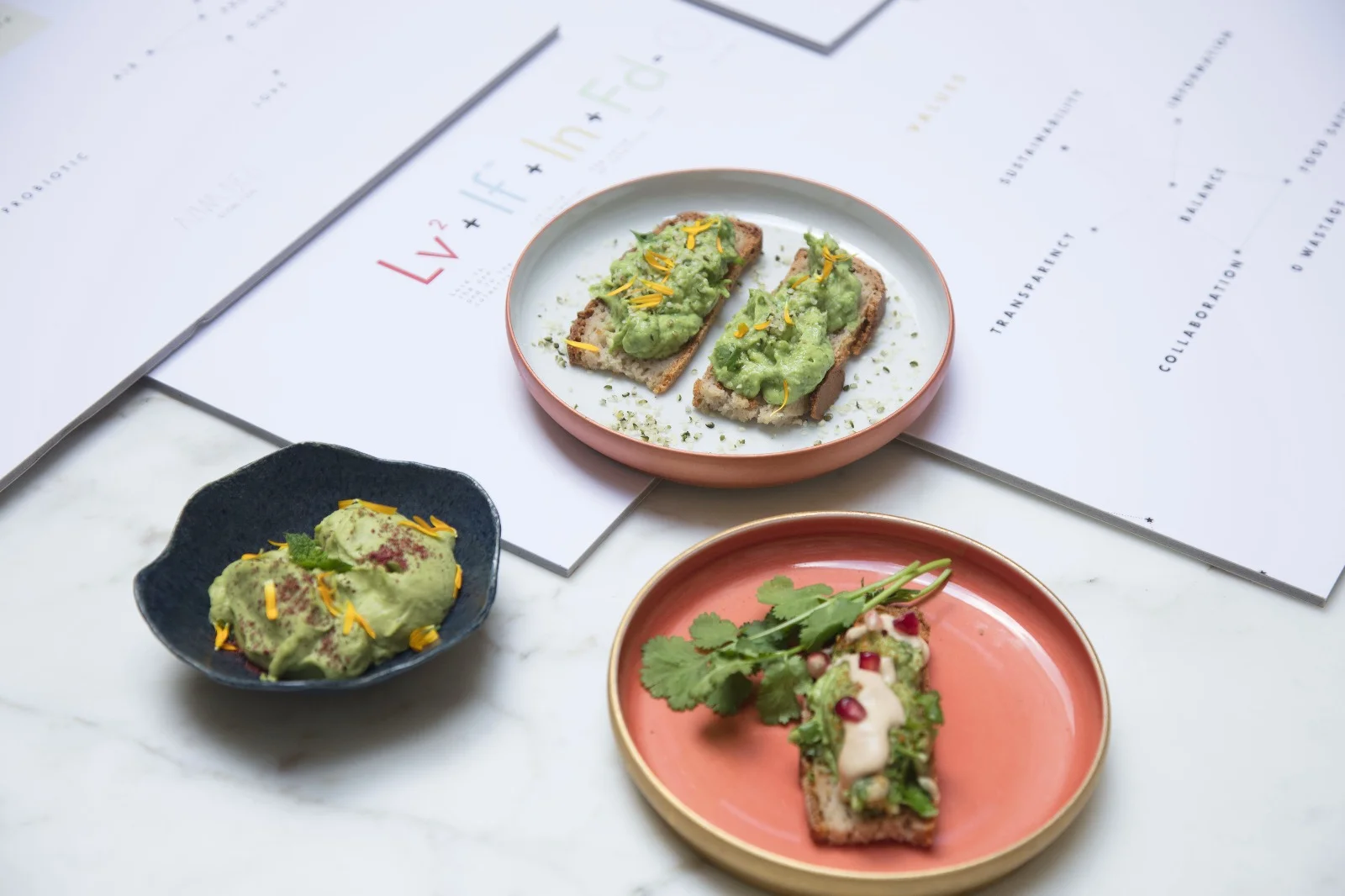 Three plates of food on a table with papers in the background. The dishes include avocado toast with toppings, and a small serving of guacamole with garnish.