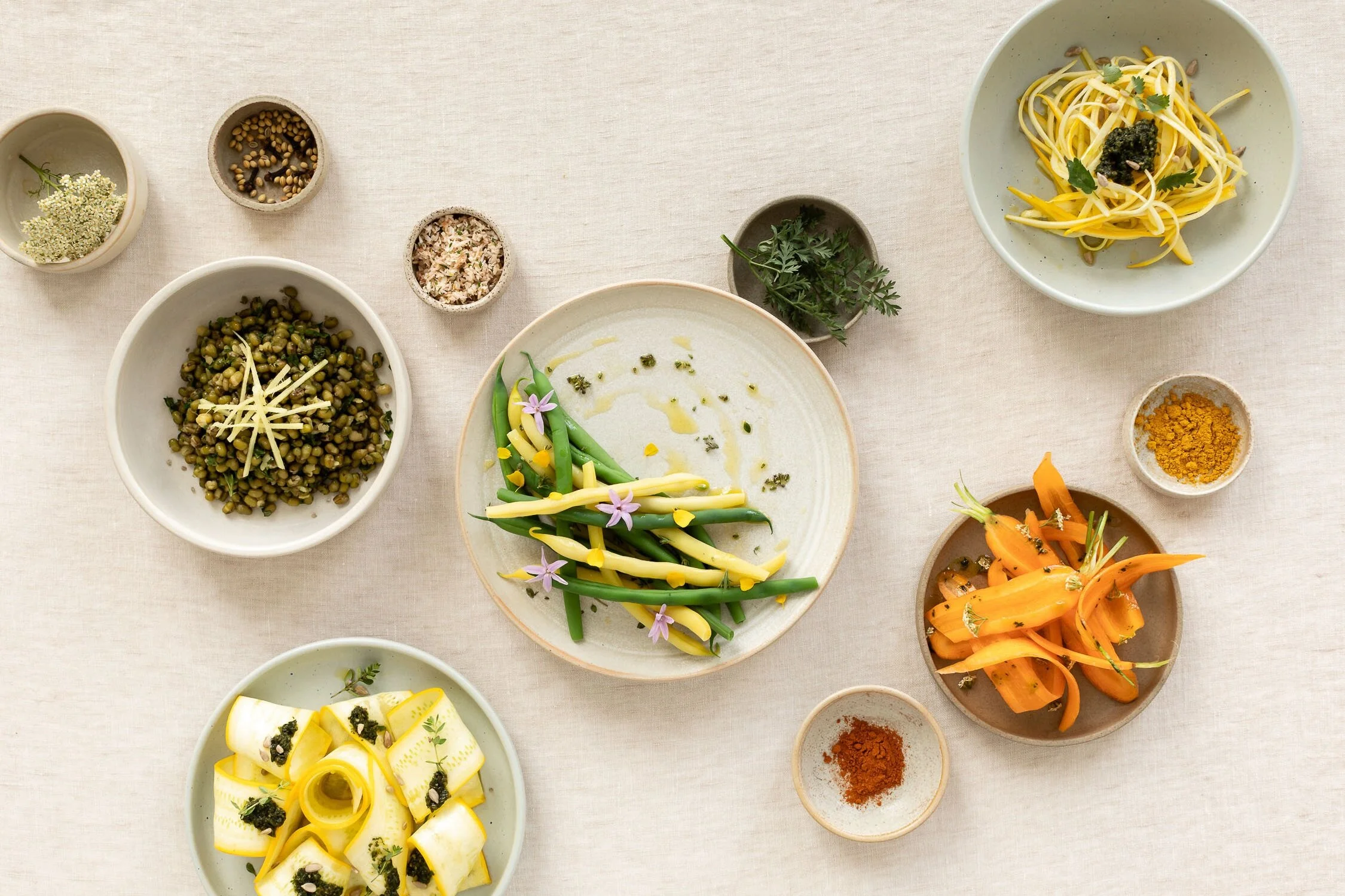 Assorted bowls containing vegetables, herbs, spices, and pasta on a beige tablecloth.