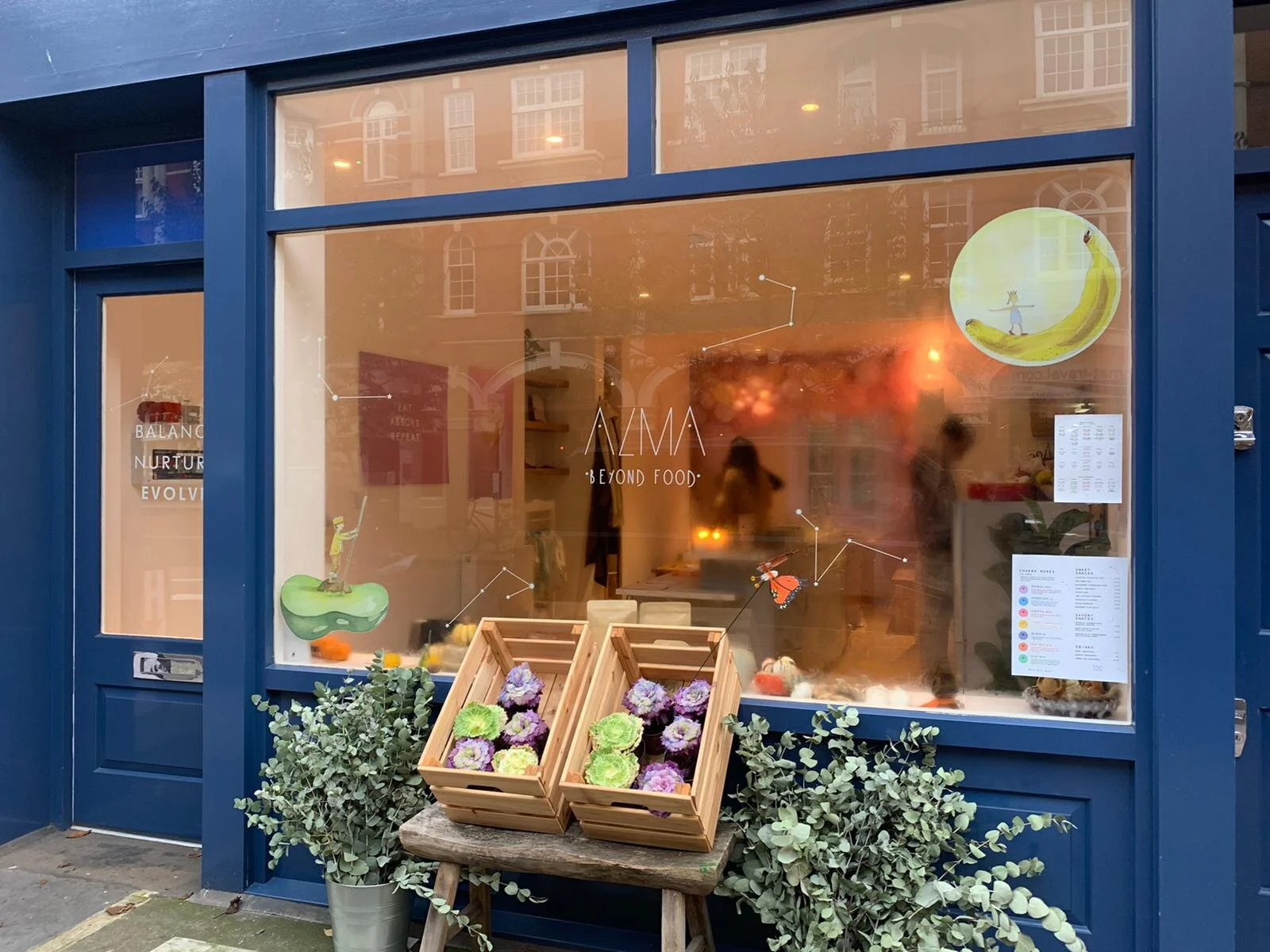 Storefront with large blue window and door, decorated with colorful drawings of animals and constellations. A wooden table outside displays purple and green flowers in boxes, flanked by two potted plants. The store's name is 'ALMA BEYOND FOOD' written on the window.