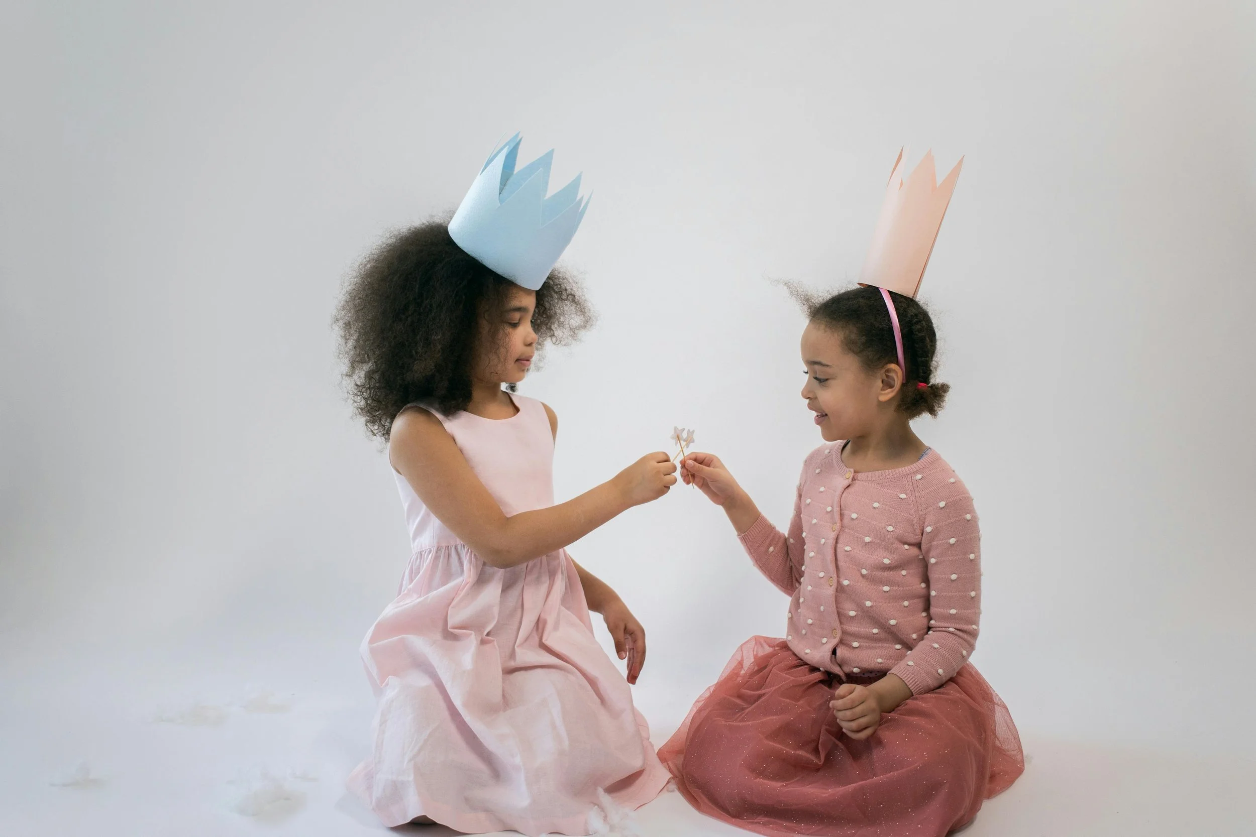 Two young girls wearing paper crowns and pink dresses sitting on the floor, playing together and exchanging a small toy, representing imagination, childhood play, and storytelling.