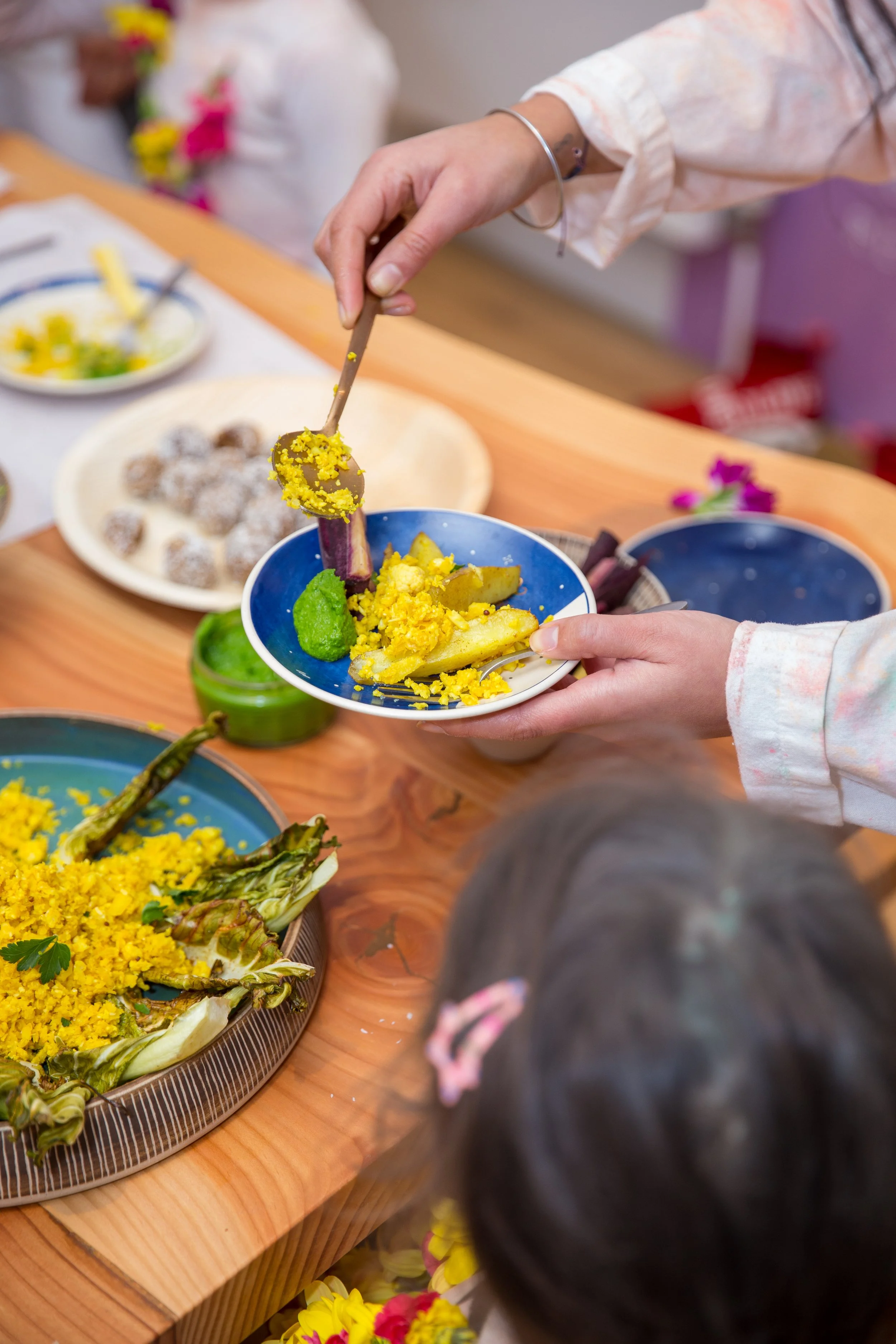 Adult serving colorful baby-led weaning meal with vegetables and textured foods onto a child’s plate at a wooden table.