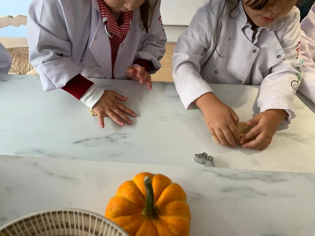 Children in chef uniforms engaging in a baking activity at a white marble table, with a pumpkin and pie cutters visible.
