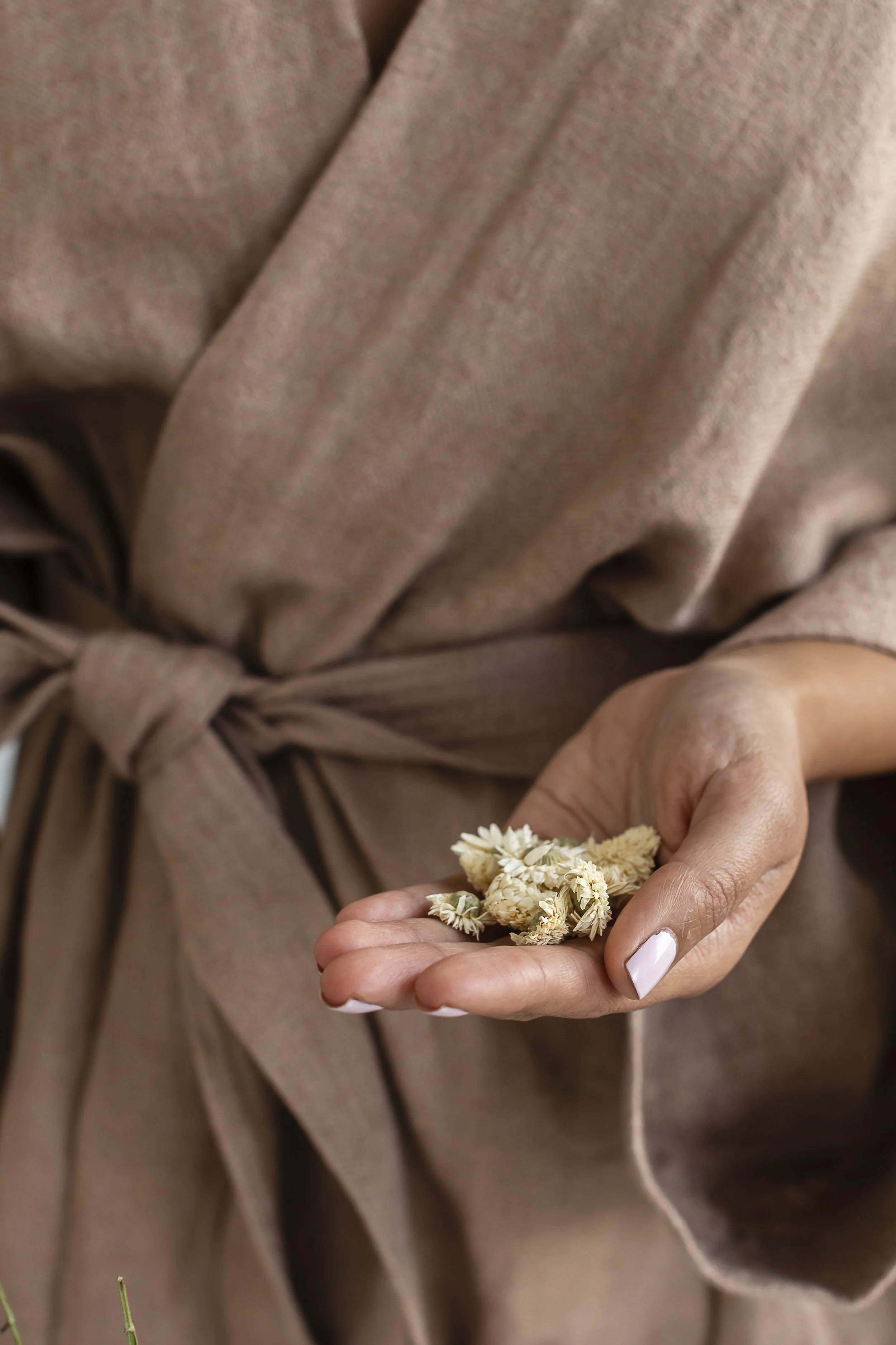 Person in a soft brown robe holding dried flowers in their hand, creating a calm and earthy wellness-inspired aesthetic.