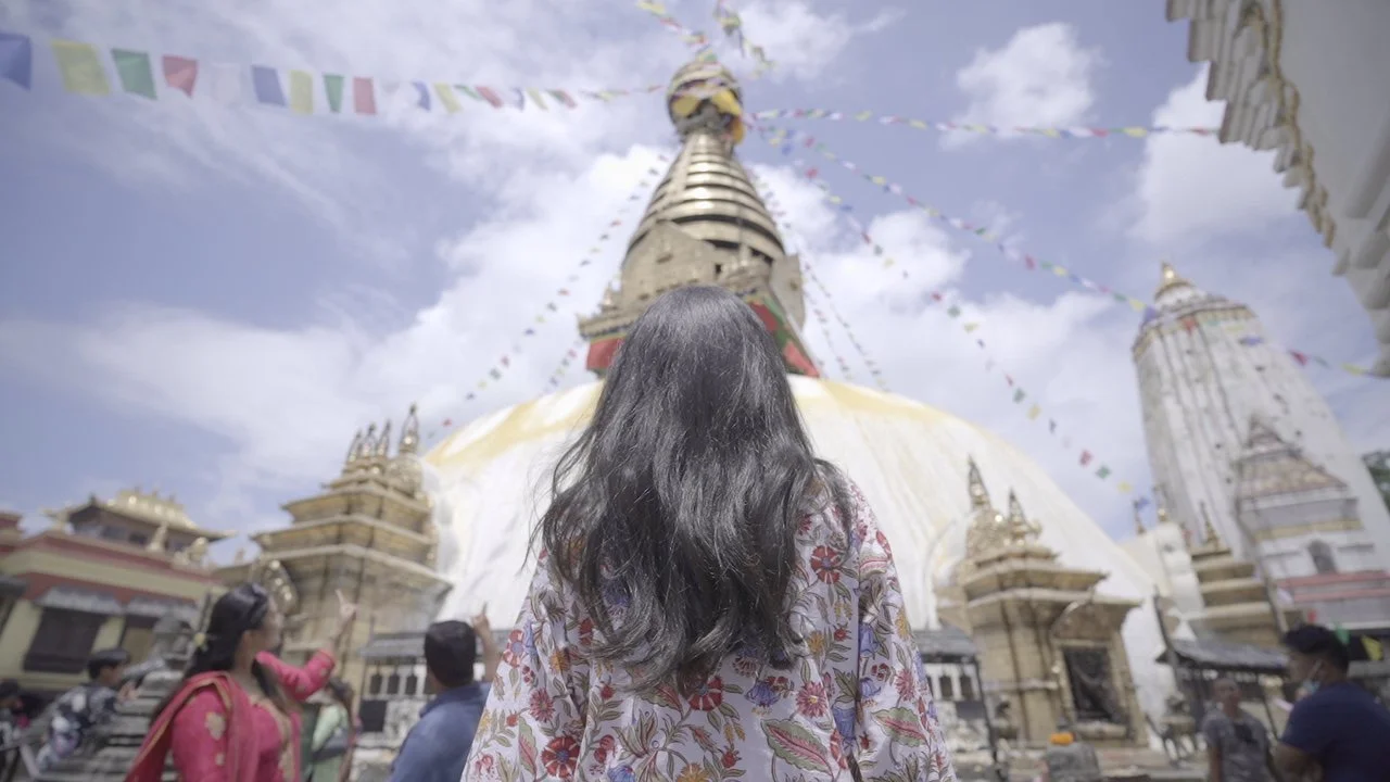 A woman with long black hair, wearing a floral patterned outfit, stands in front of a large Buddhist stupa adorned with prayer flags, in a busy temple courtyard of a Nepalese temple.