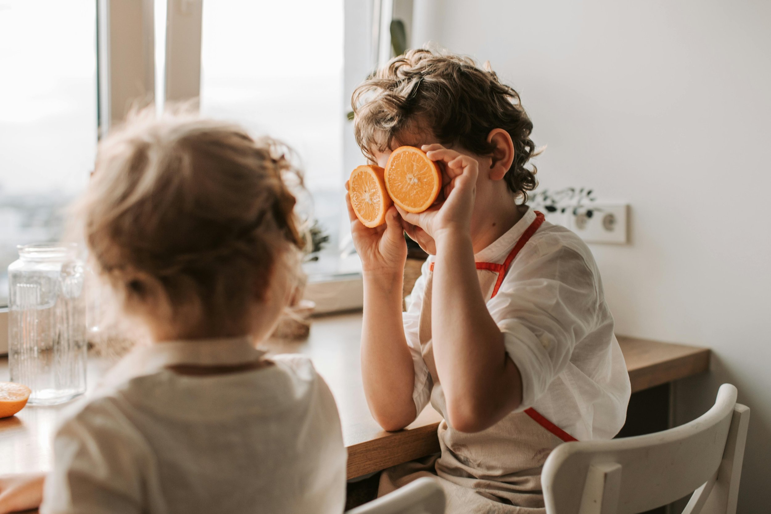 A young child sits at a kitchen table holding two orange slices up to their eyes, playfully pretending they are glasses, while another child sits nearby watching.