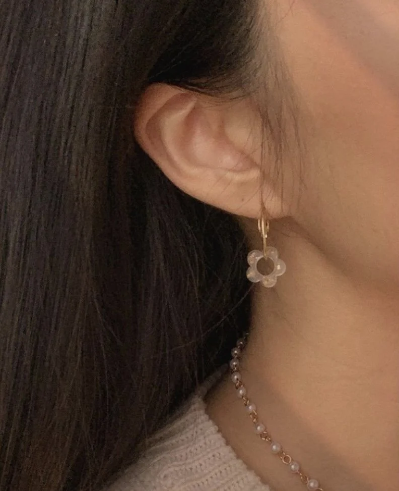 Close-up shot of a person wearing a gold hoop earring with a clear resin flower charm alongside a delicate pearl chain necklace.