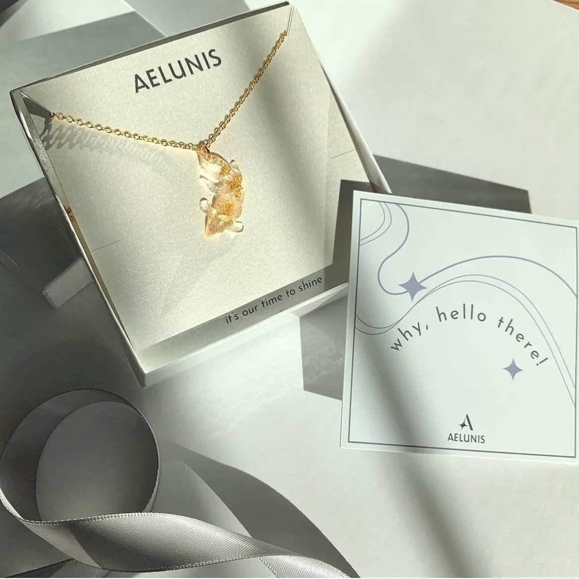 Packaged gold necklace with a clear and gold flake resin pendant displayed in an Aelunis brand box alongside a brand insert card and white ribbon.