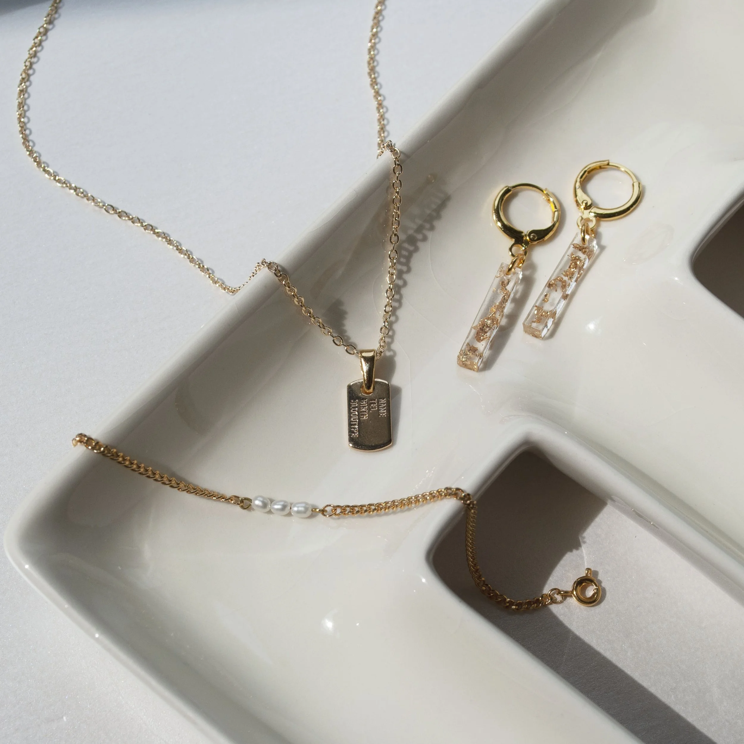 Gold jewelry set featuring a dog tag pendant necklace, a thin chain bracelet with tiny pearl accents, and rectangular clear resin drop earrings with gold flake details, resting on a white ceramic tray.