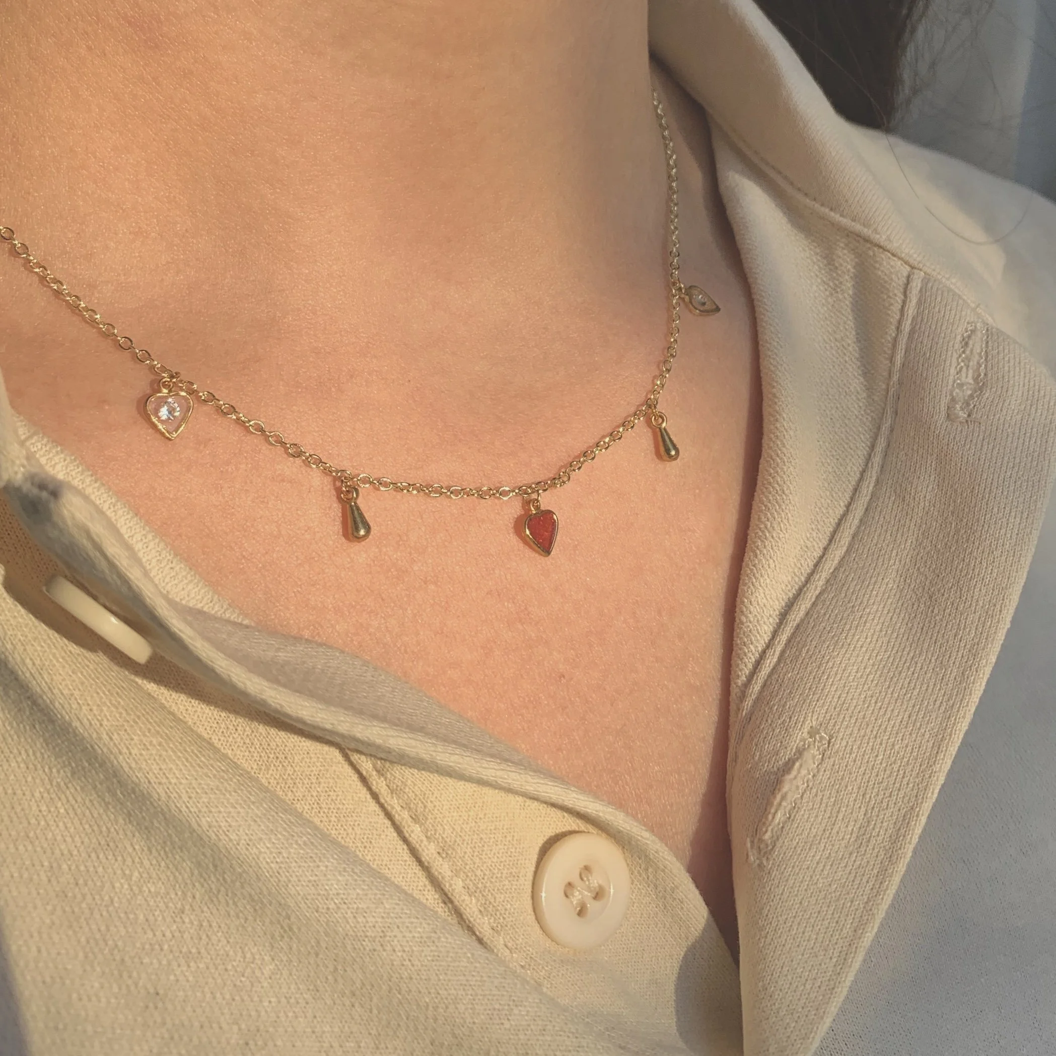 Close-up shot of a person wearing a delicate gold charm necklace with small drop charms and heart pendants, bathed in warm sunlight.