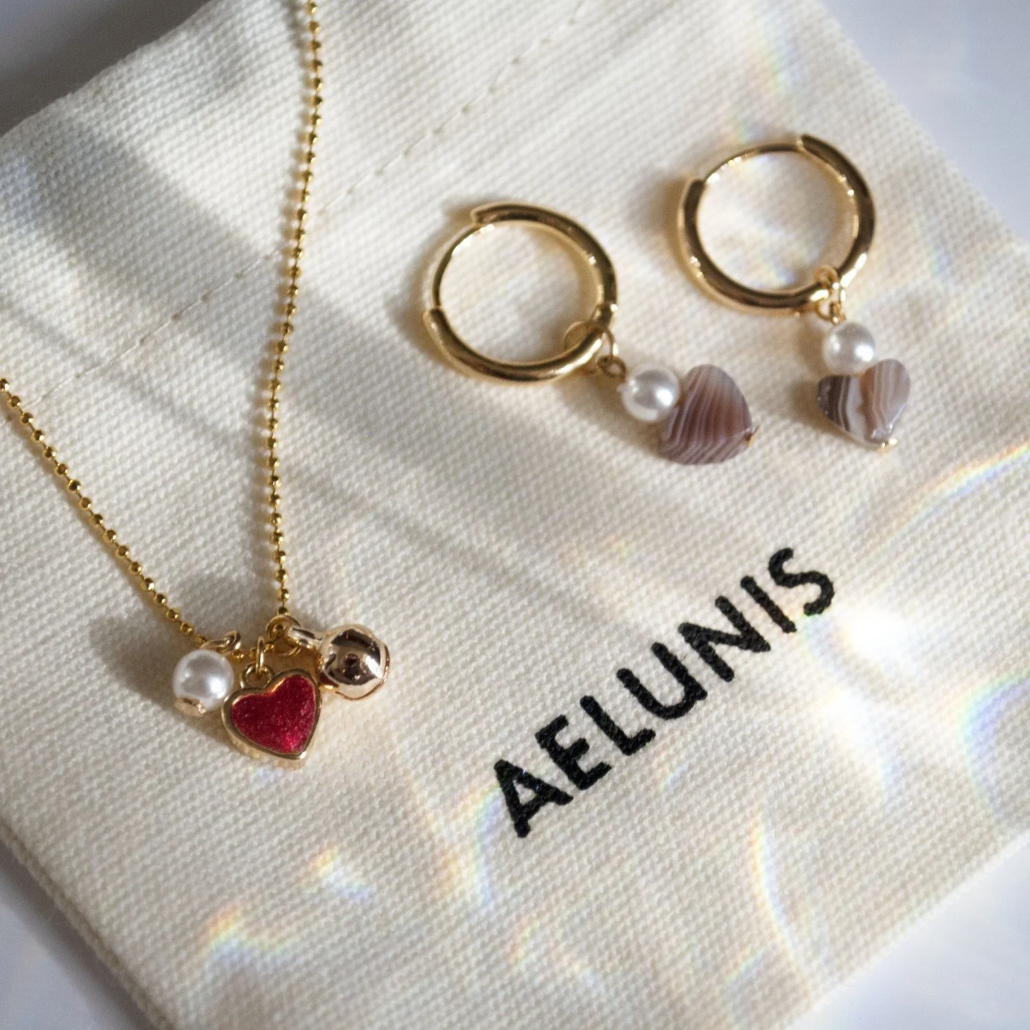 Gold ball-chain charm necklace and heart hoop earrings resting on an Aelunis cream fabric pouch with soft sunlight reflection.