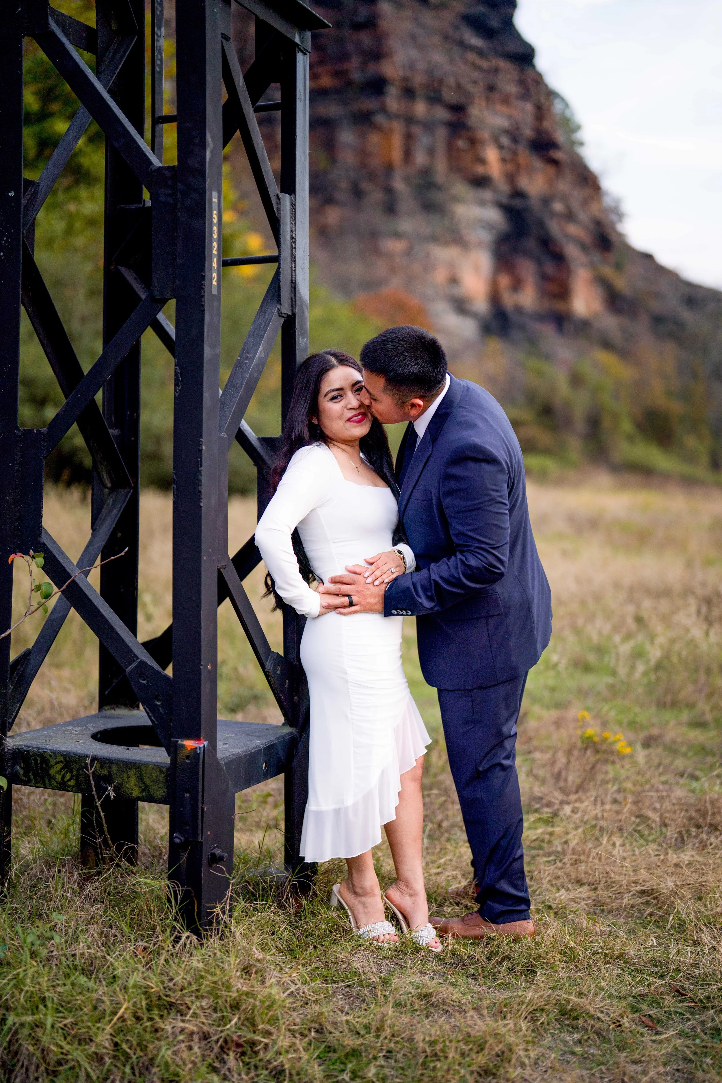 A couple dressed formally, with the man in a navy suit and the woman in a white dress, sharing an intimate moment outdoors near a metal structure with a rocky hill in the background.