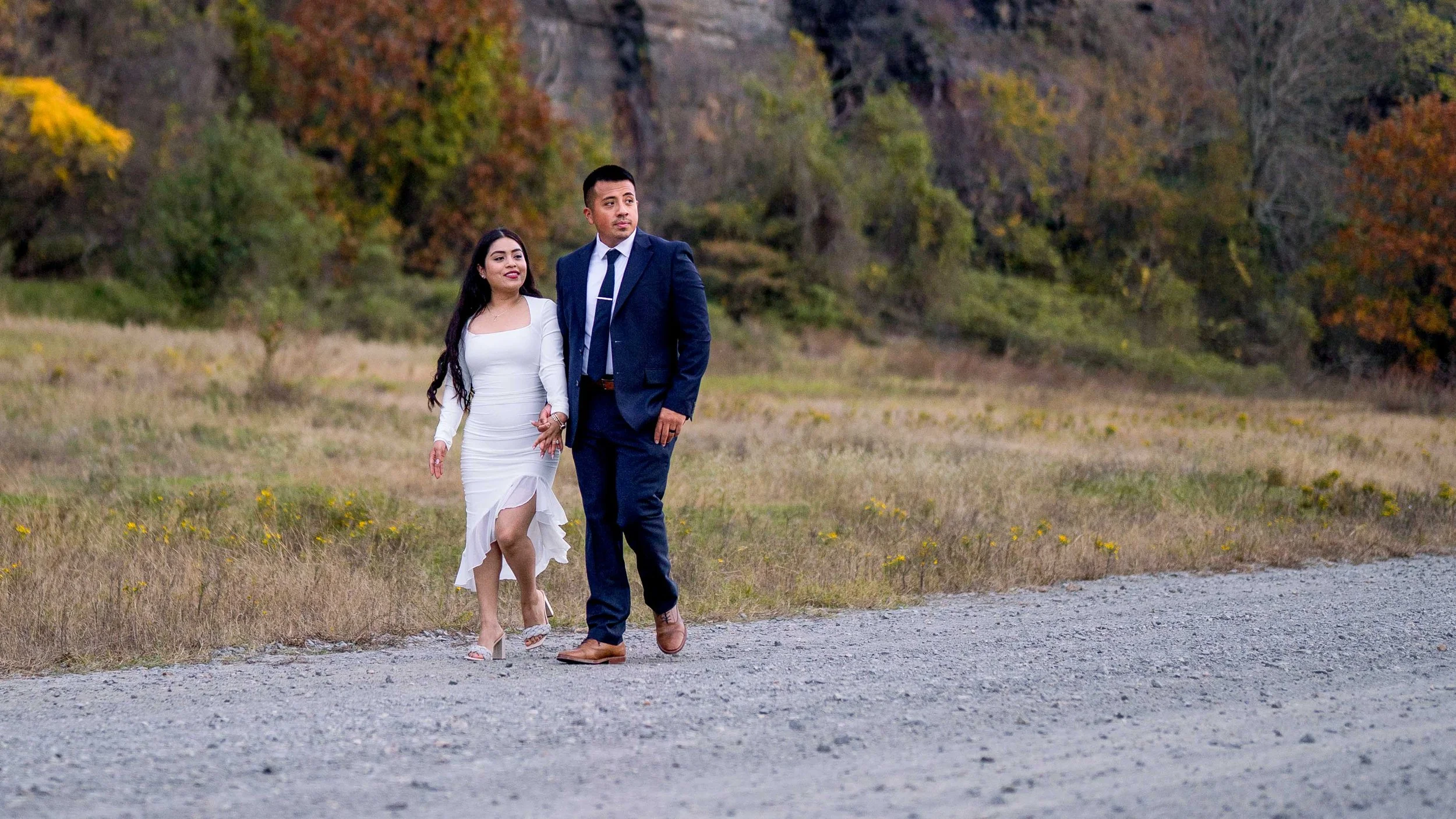 A couple dressed in formal attire walking in an open field with trees showing fall colors in the background.