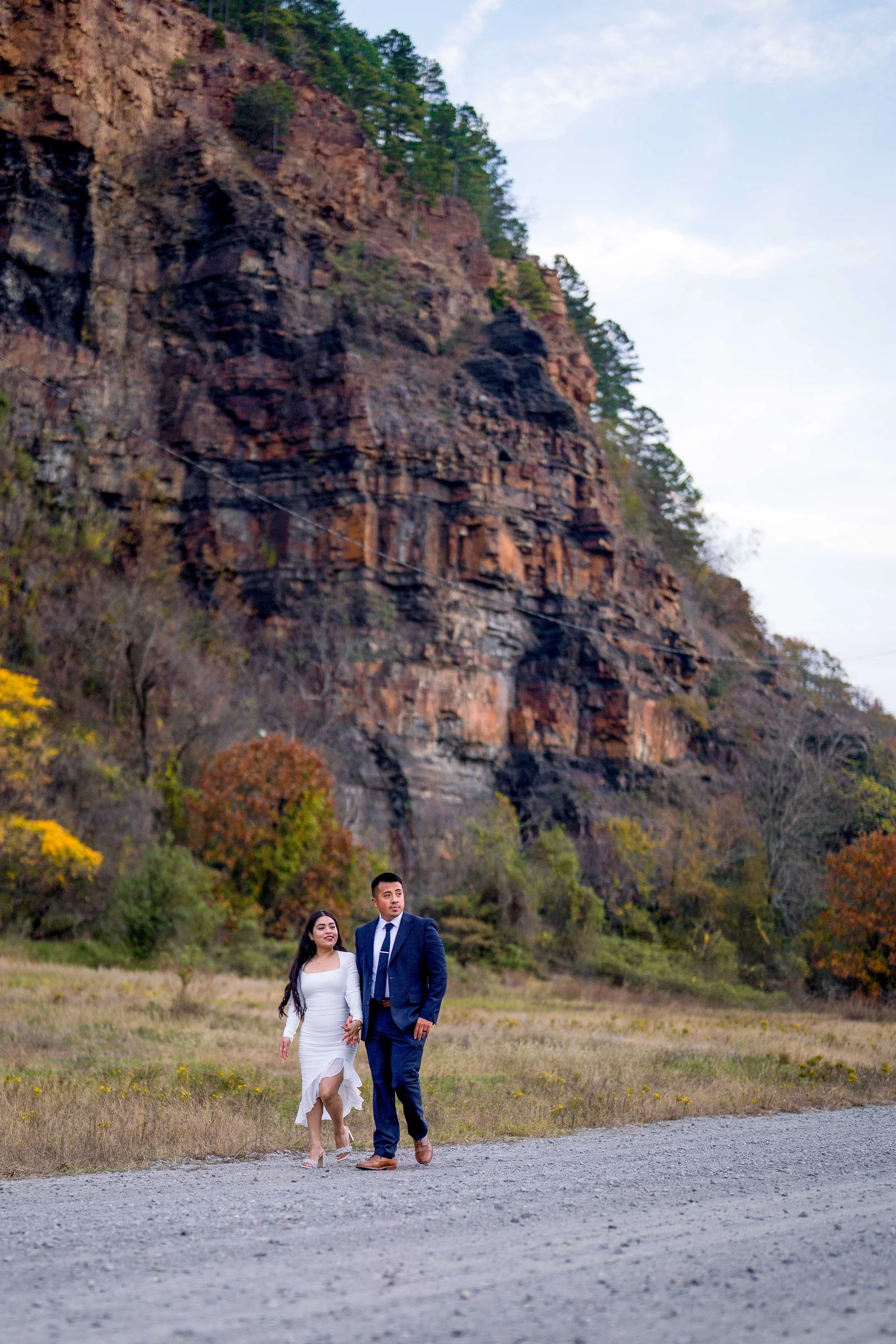 A couple dressed in formal attire walking outdoors near a rocky cliff and autumn-colored trees.
