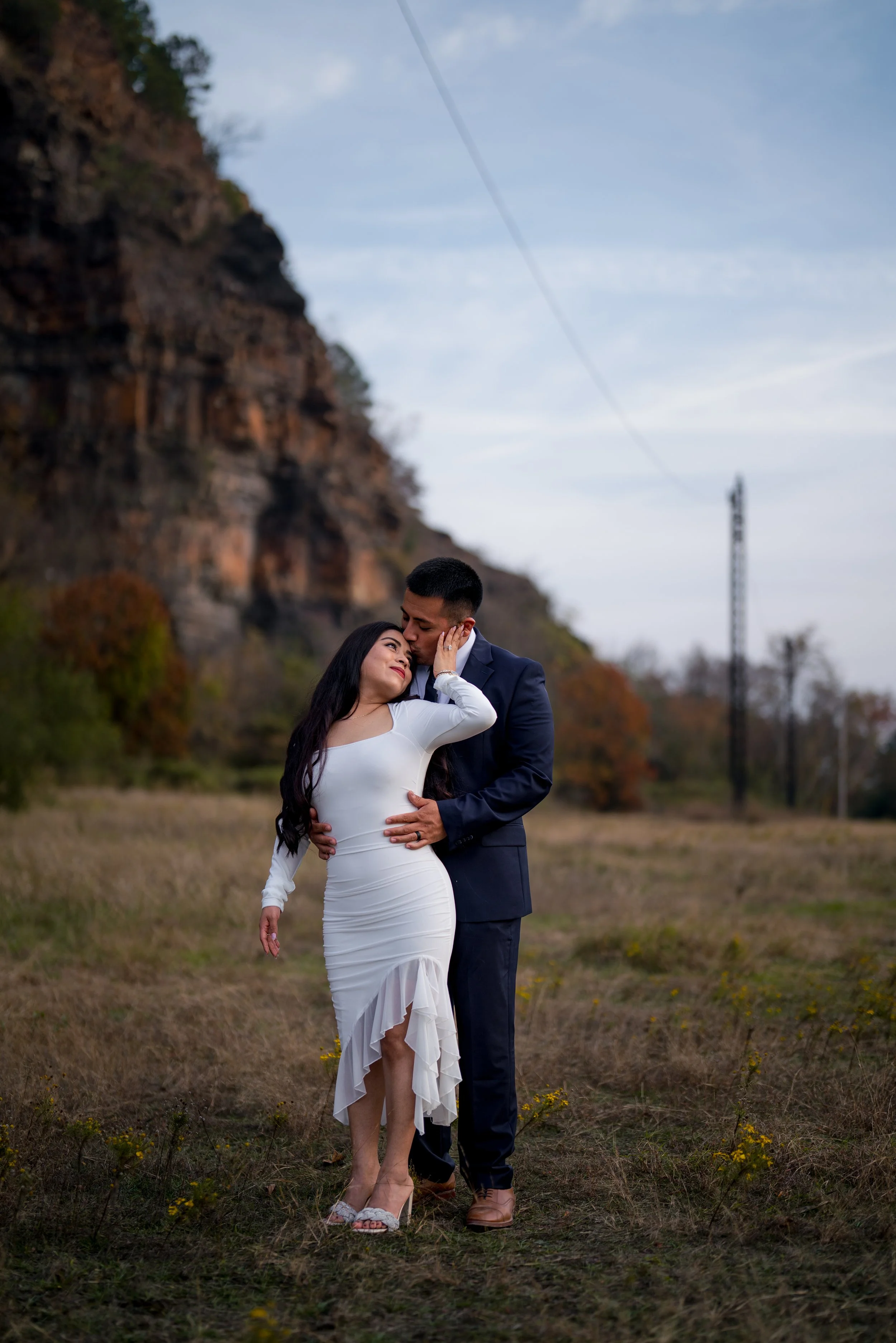 A romantic couple dressed in formal attire sharing a tender moment in an outdoor field during dusk, with a rocky hillside and power lines in the background.