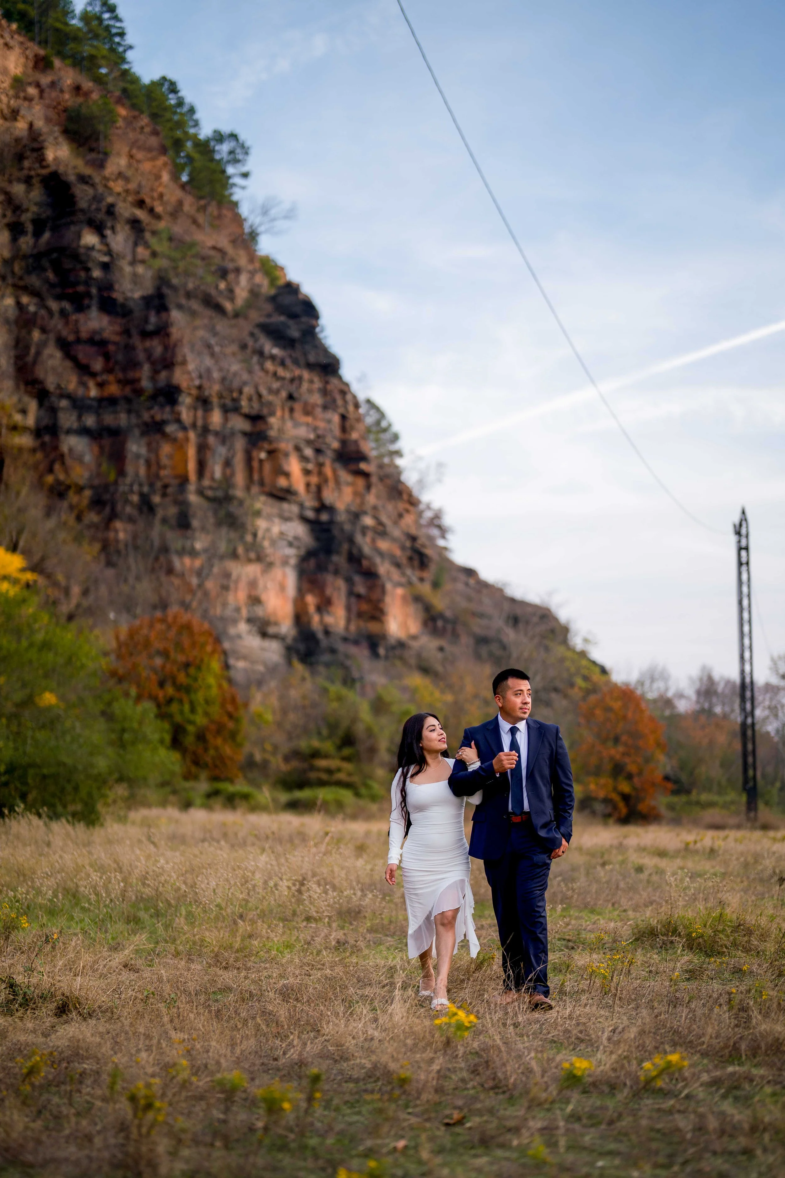 A woman and man walking together in a grassy field with trees and a rocky hill in the background, during daytime.