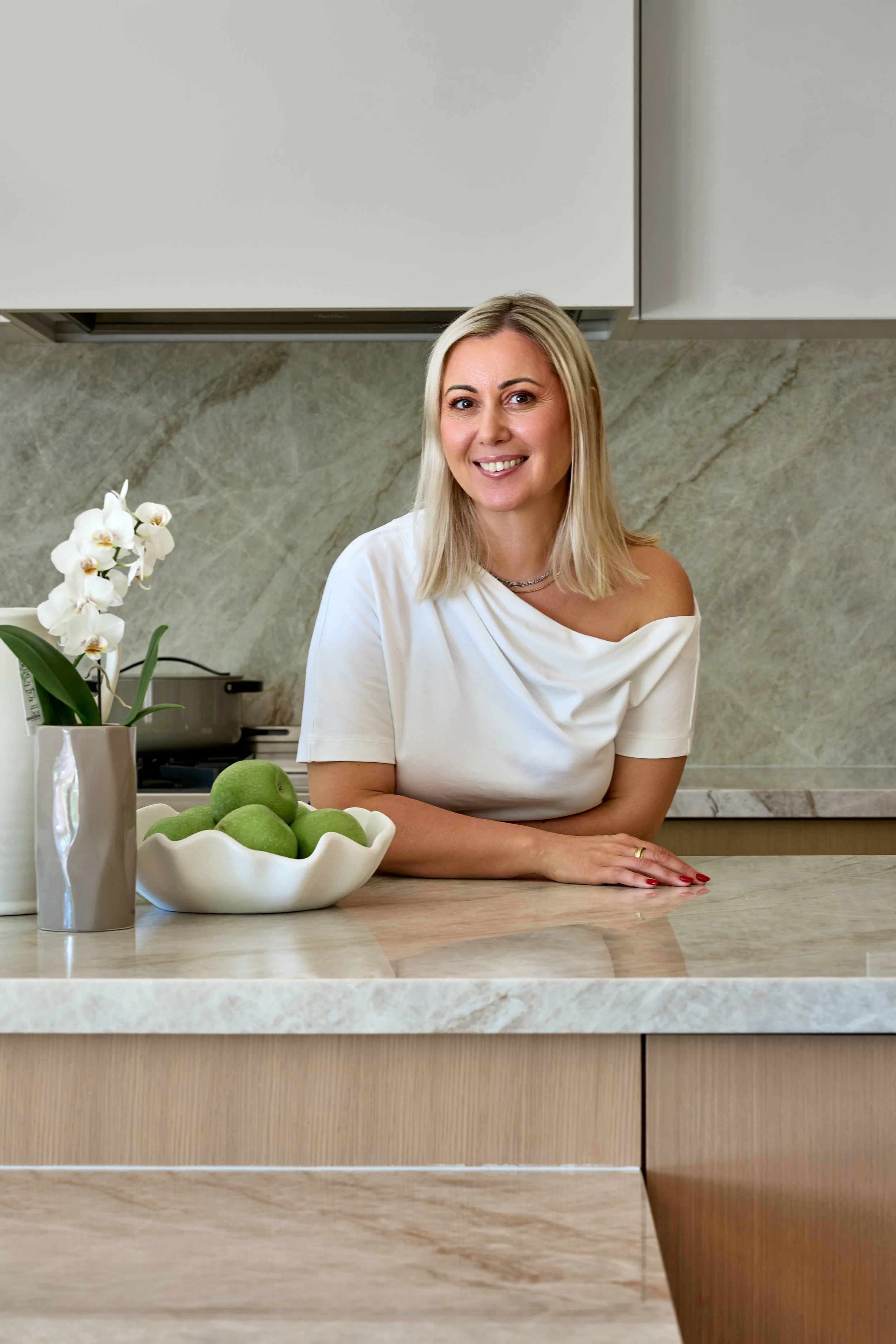 A woman with blonde hair smiling and sitting at a kitchen counter, with a bowl of green apples and a white orchid plant nearby.