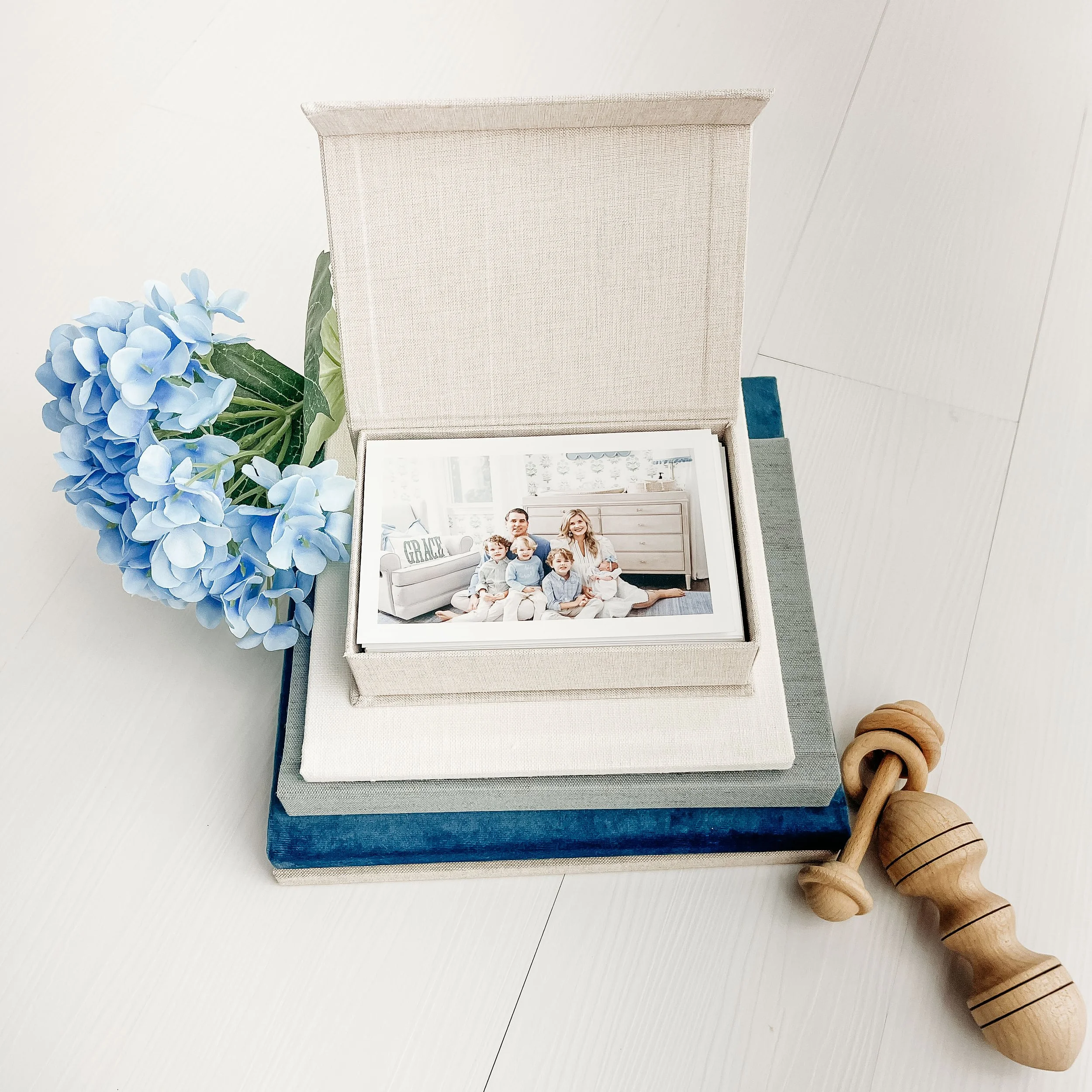 Family photo in fabric box, blue hydrangea, books, wooden rattle on white floor.