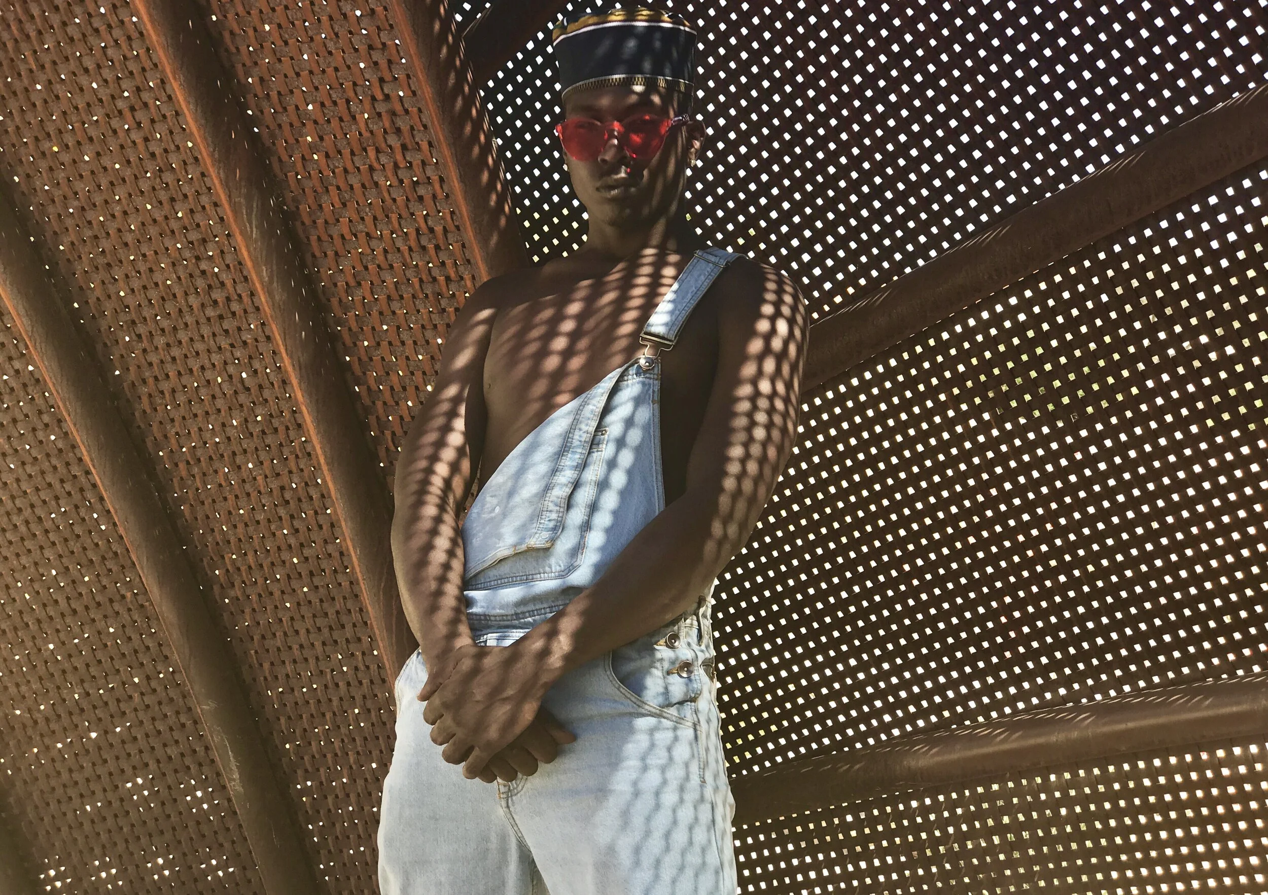 A shirtless young man wearing red sunglasses, a black cap, and light blue denim overalls, standing with hands clasped in front, against a perforated wooden structure with sunlight creating a pattern of light and shadow.