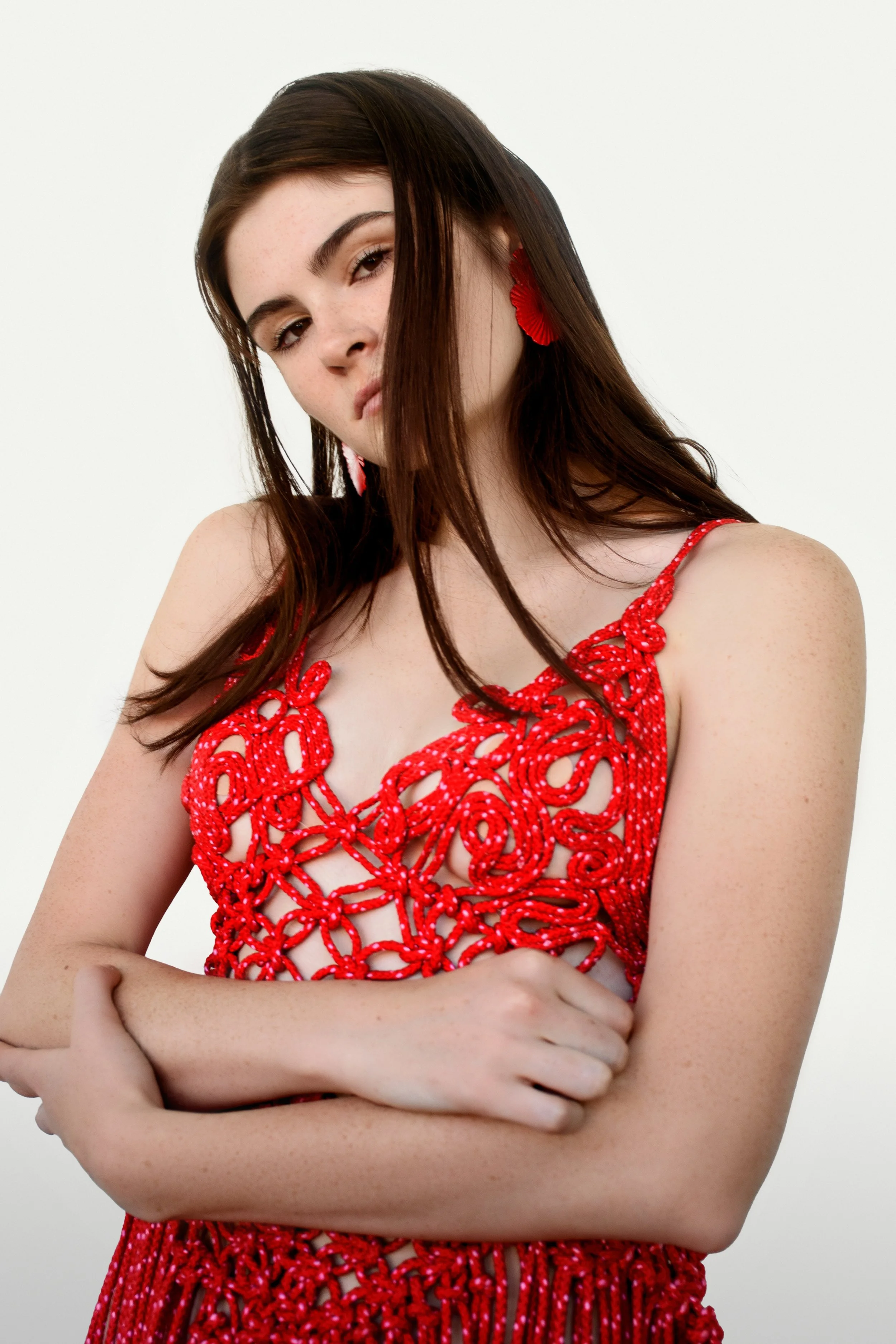 A portrait of a model in a bold red macramé dress, featuring intricate knotting and cut-out details. She pairs the look with her piercing gaze. The clean, minimal backdrop emphasizes the sculptural, high-fashion aesthetic. Photo by Naomy Quiñones.